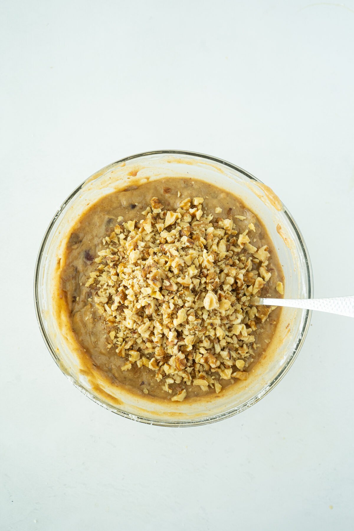 A glass bowl filled with banana bread batter topped with chopped walnuts, with a metal spoon resting inside, on a white surface.
