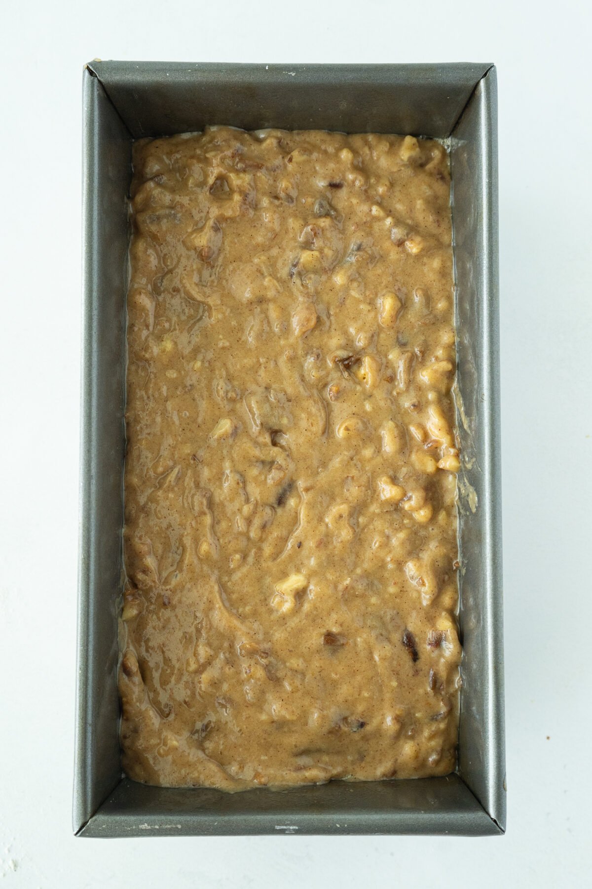 A metal loaf pan filled with unbaked banana bread batter, containing visible chunks of banana and nuts, sits on a white surface.