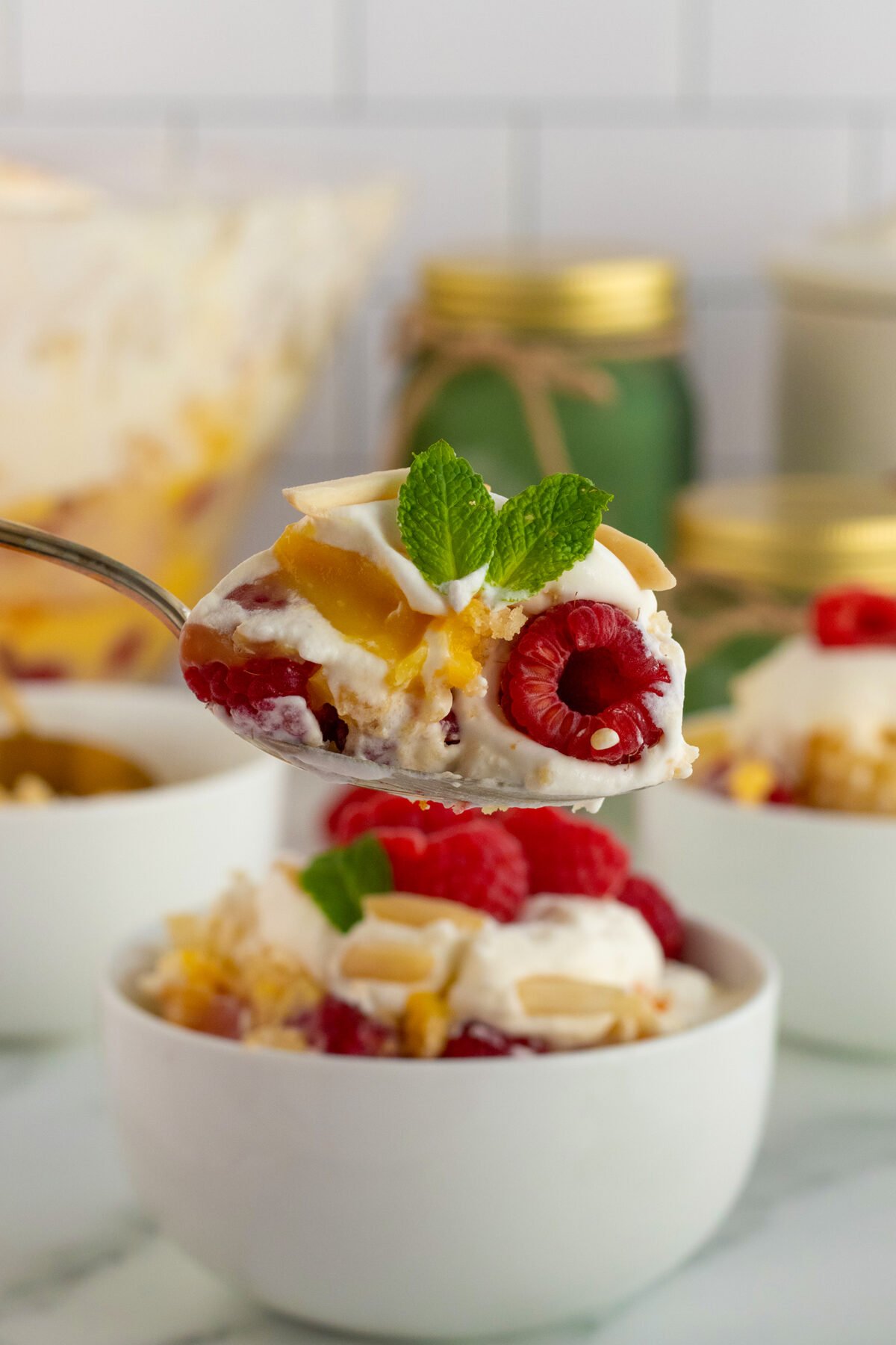 A spoonful of trifle with whipped cream, raspberries, almonds, and mint is held above a white bowl filled with the same dessert. There are jars and another bowl blurred in the background.