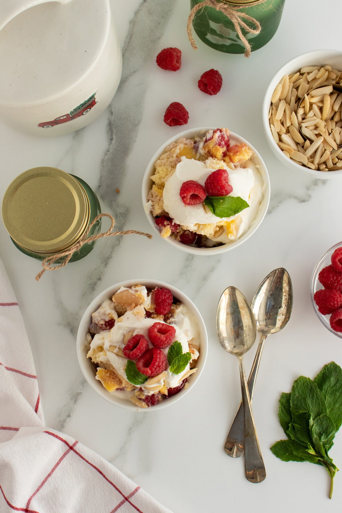 Two bowls of dessert topped with whipped cream, fresh raspberries, and mint leaves sit on a marble surface beside two spoons, a bowl of sliced almonds, a bowl of raspberries, and green jars.