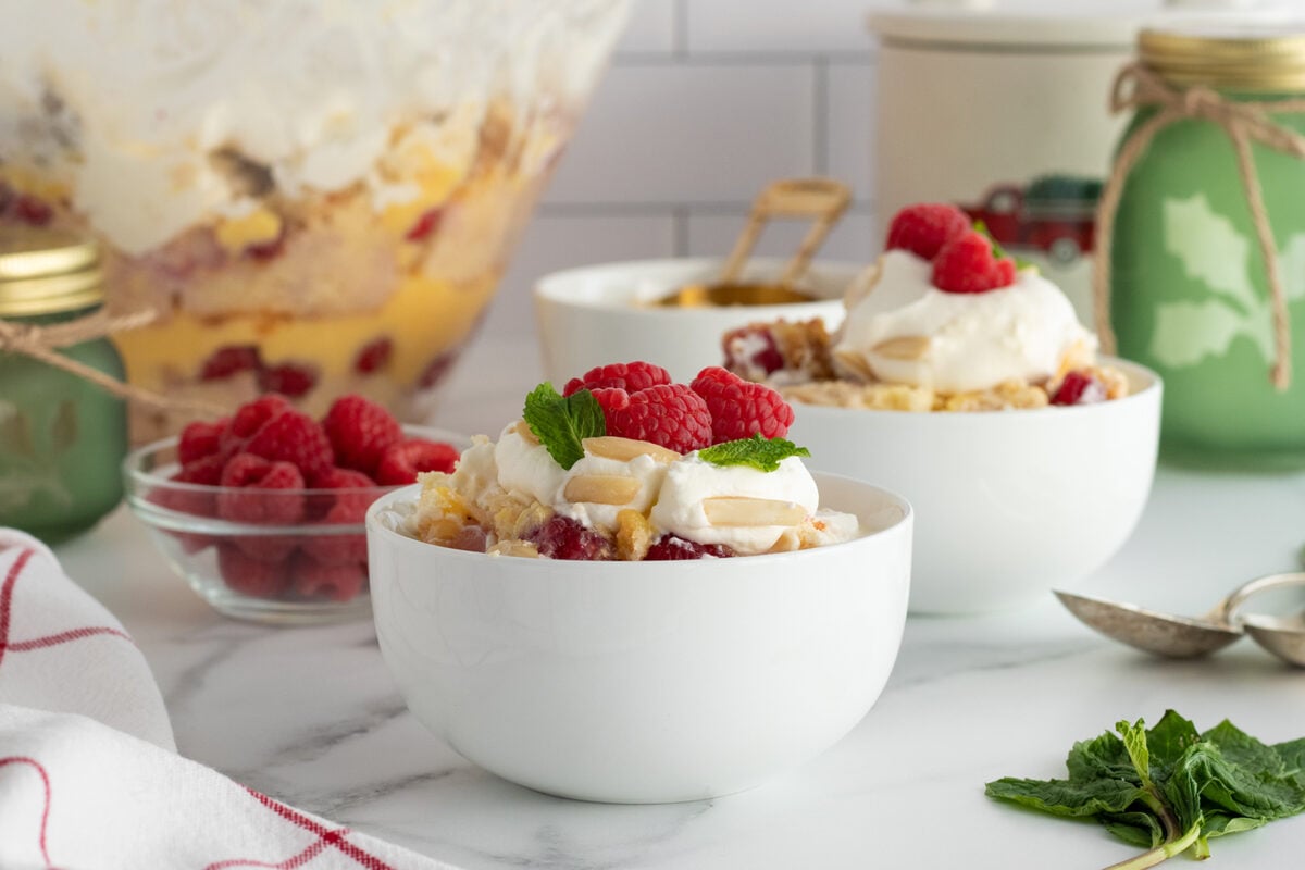 Two white bowls filled with a layered dessert topped with whipped cream, fresh raspberries, slivered almonds, and mint leaves. Fresh raspberries, green jars, and a glass bowl of dessert are in the background.