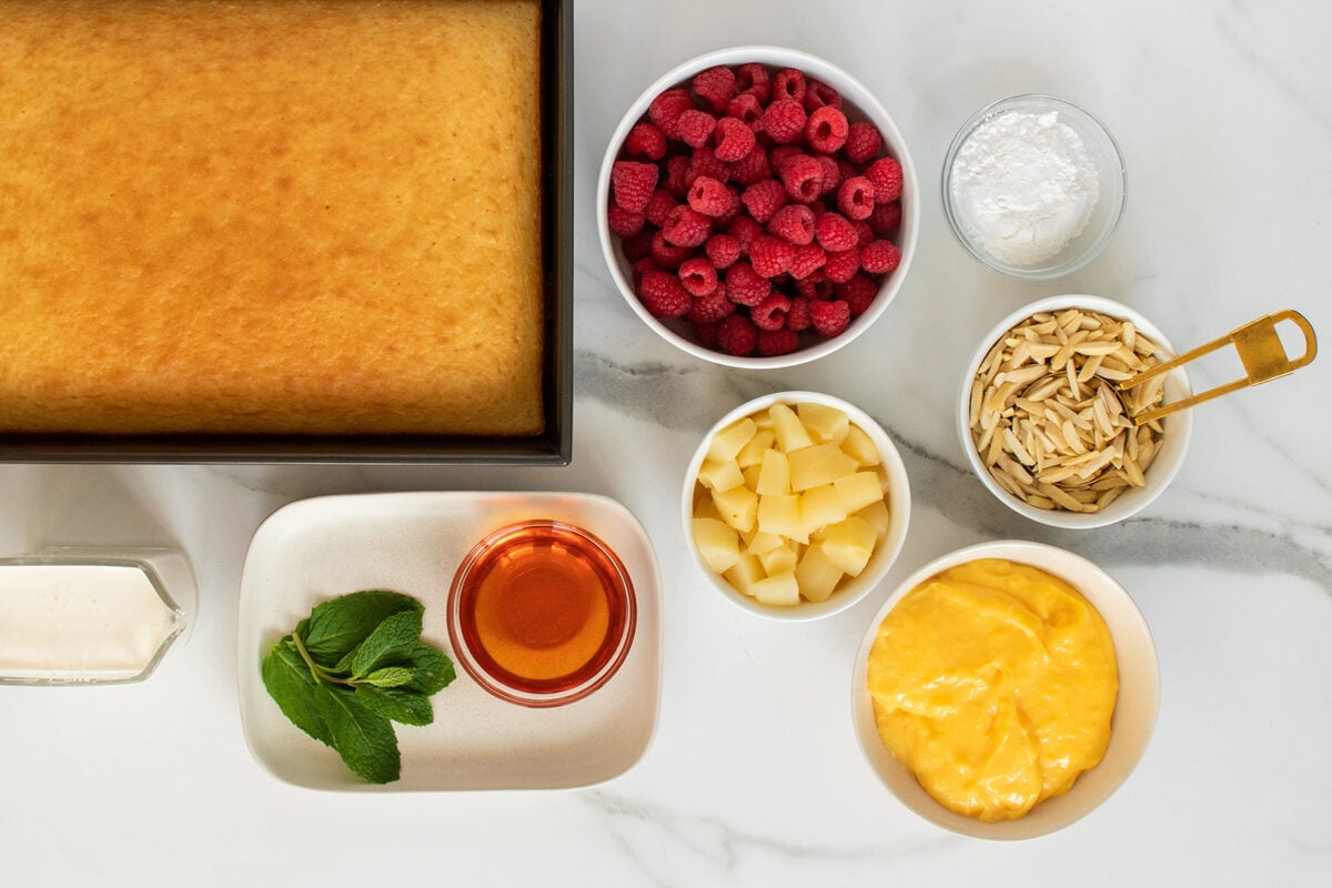 A rectangular sponge cake in a pan is surrounded by bowls of raspberries, powdered sugar, slivered almonds, pineapple chunks, yellow custard, a glass of syrup, a plate with mint leaves, and a small pitcher of cream on a marble surface.