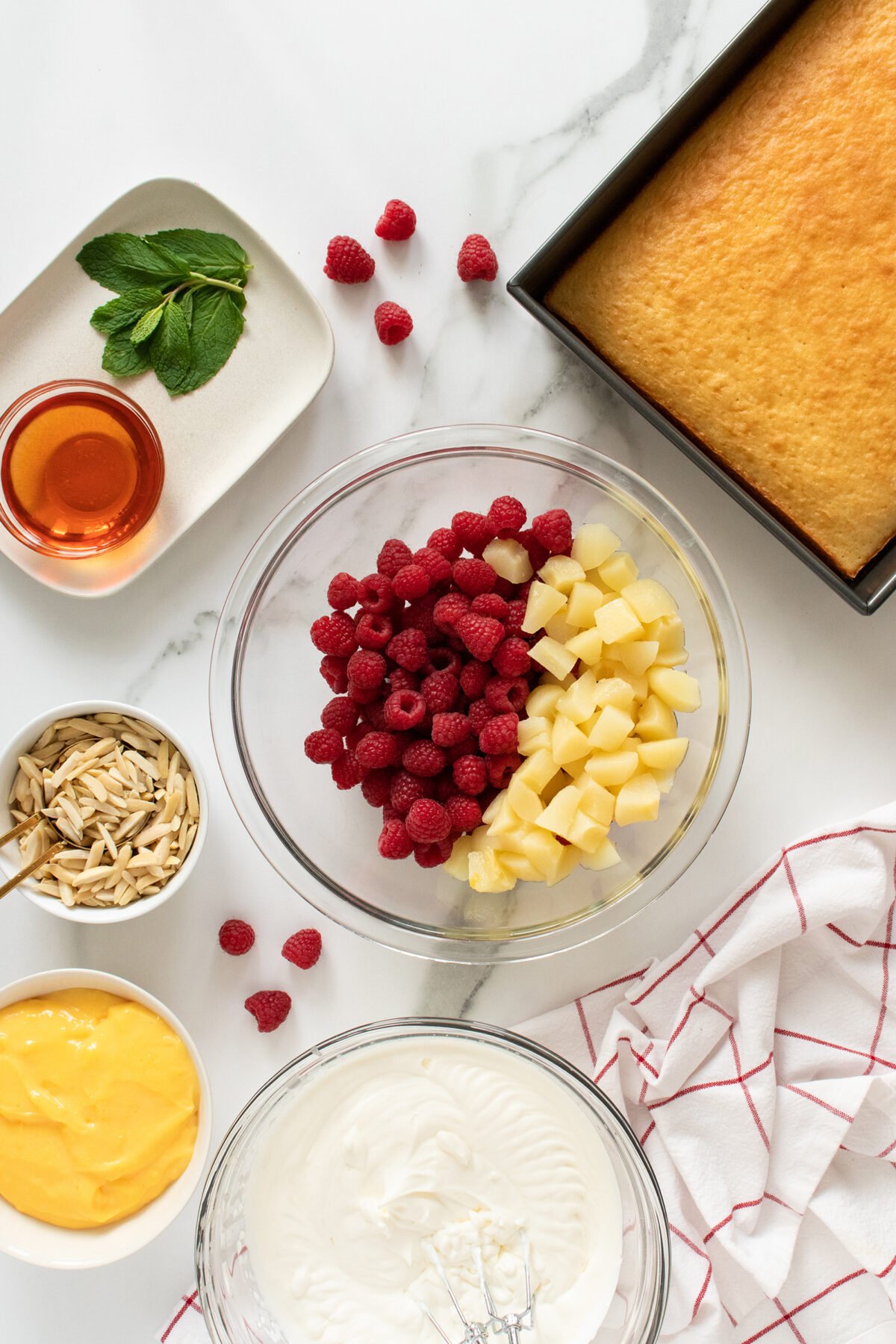 A top view of ingredients for a dessert: a bowl with raspberries and pineapple, a baked cake in a pan, whipped cream, yellow custard, slivered almonds, honey, and mint leaves on a marble surface with a red-checkered towel.