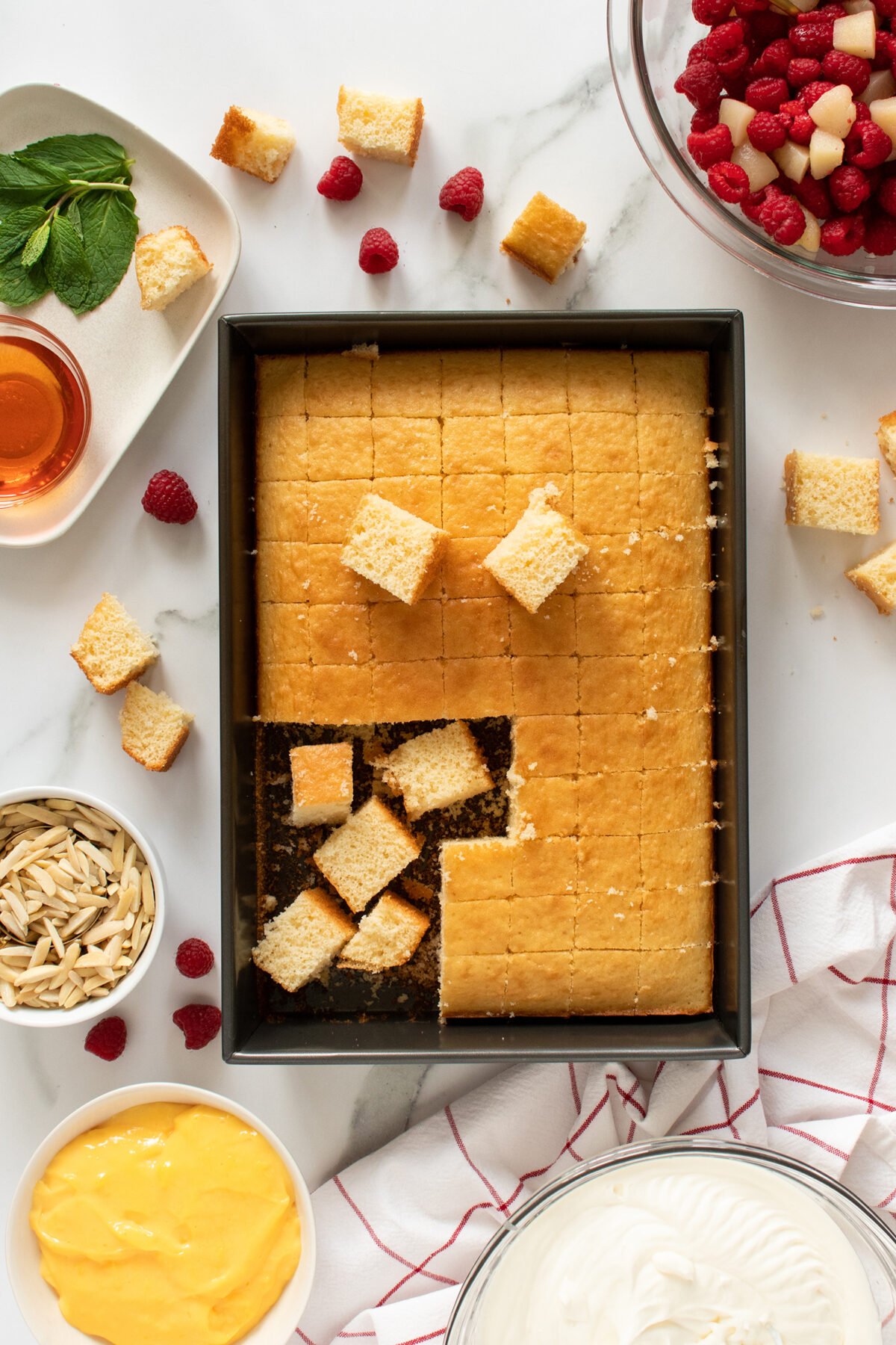 A rectangular pan of sliced yellow cake sits on a counter with some pieces removed. Surrounding it are bowls of whipped cream, custard, slivered almonds, and a bowl of mixed berries. A plate with mint, honey, and raspberries is nearby.