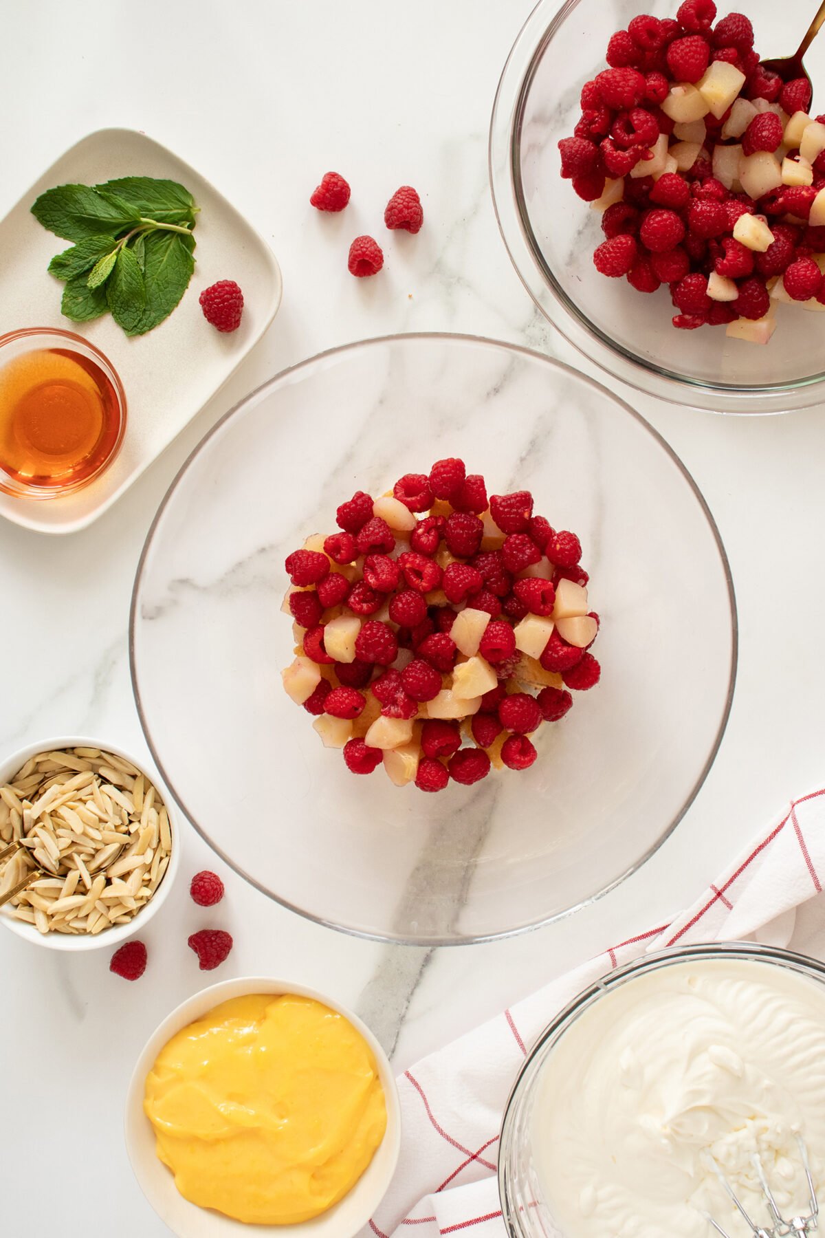 A glass bowl of raspberries and diced pears sits on a white countertop, surrounded by bowls of sliced almonds, yellow custard, whipped cream, a plate of mint leaves and honey, and a bowl of more raspberries and pears.