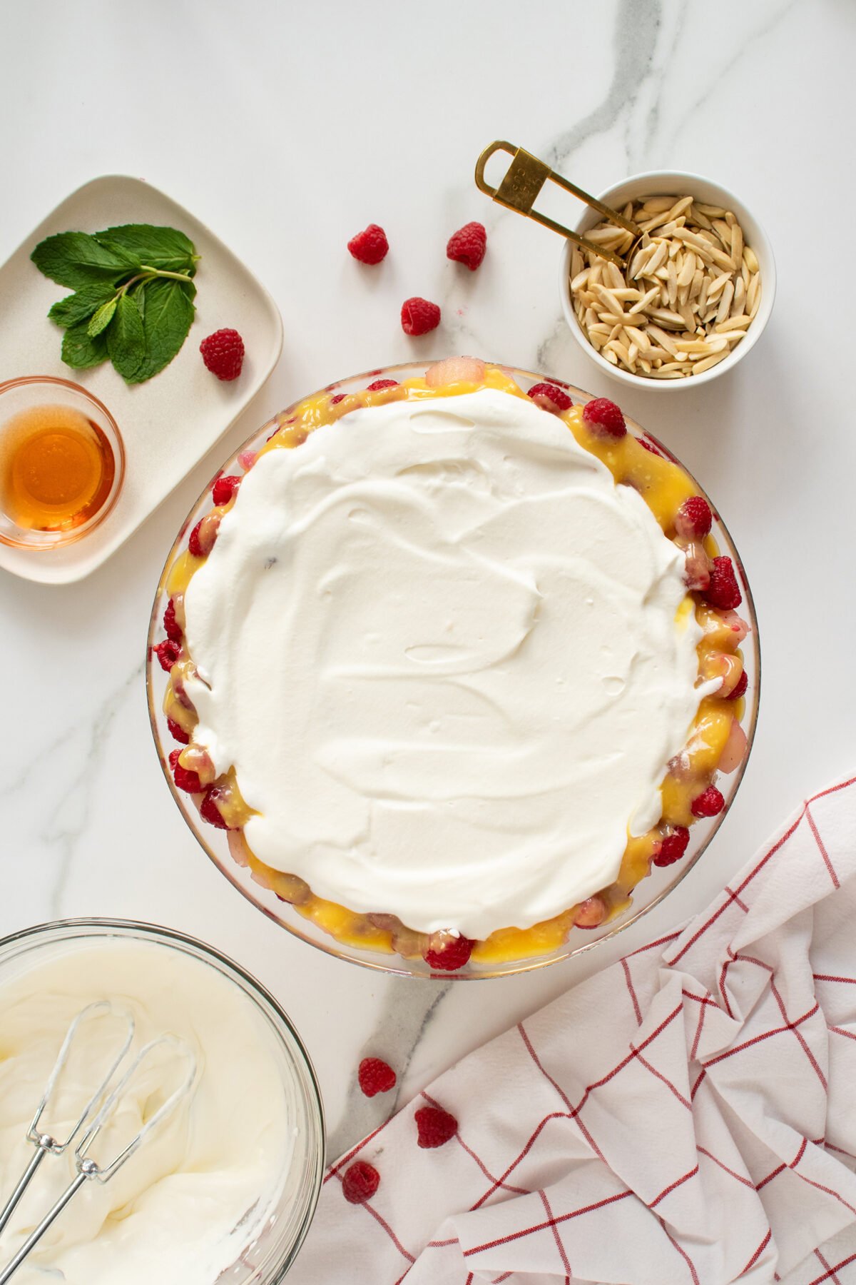 Overhead view of a trifle dessert topped with whipped cream, surrounded by raspberries, a bowl of slivered almonds, a small dish of honey, fresh mint leaves, a bowl of whipped cream, and a red-and-white kitchen towel.