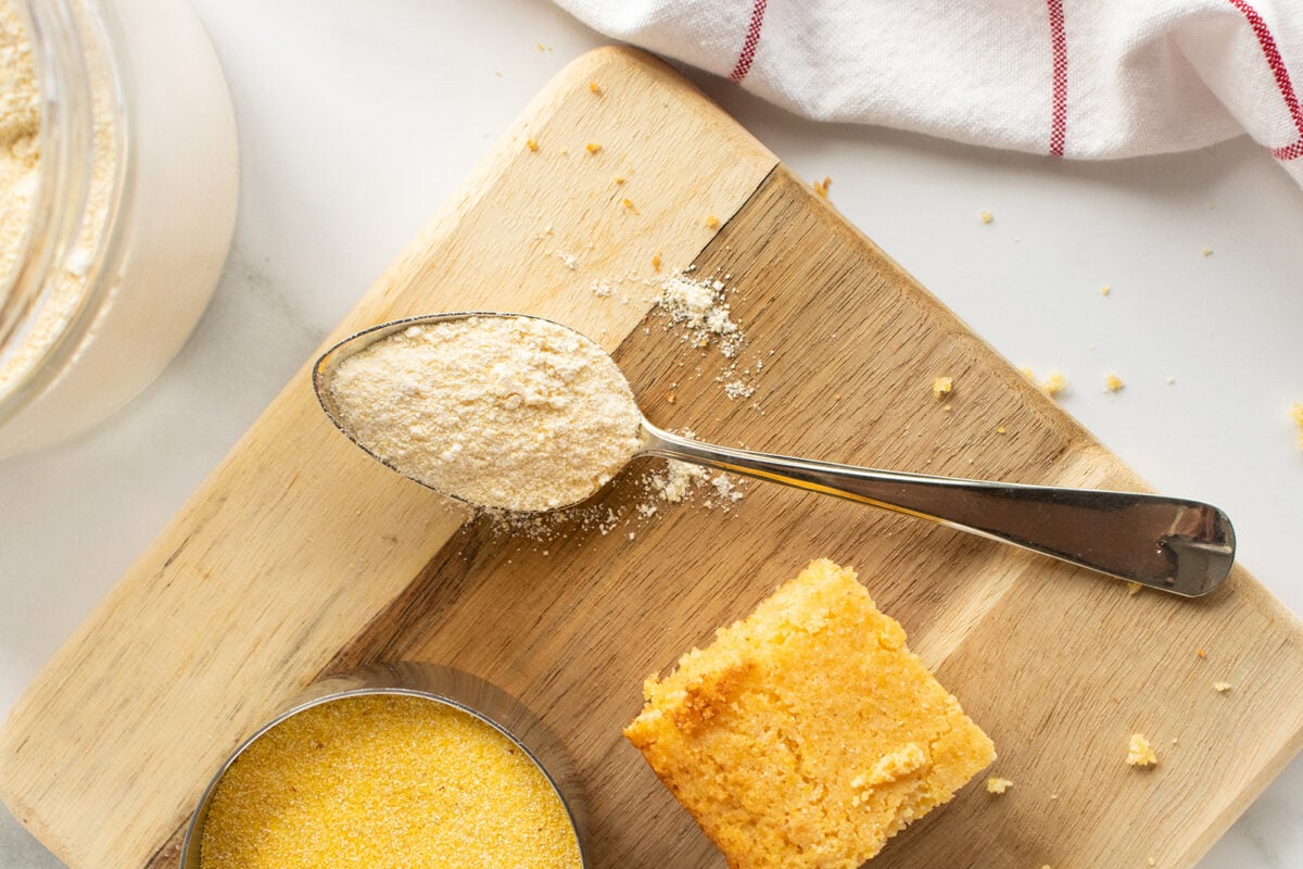 A metal spoon with cornmeal rests on a wooden cutting board next to a square piece of cornbread, a small dish of cornmeal, and a white towel with a red stripe. Cornmeal crumbs are scattered around.