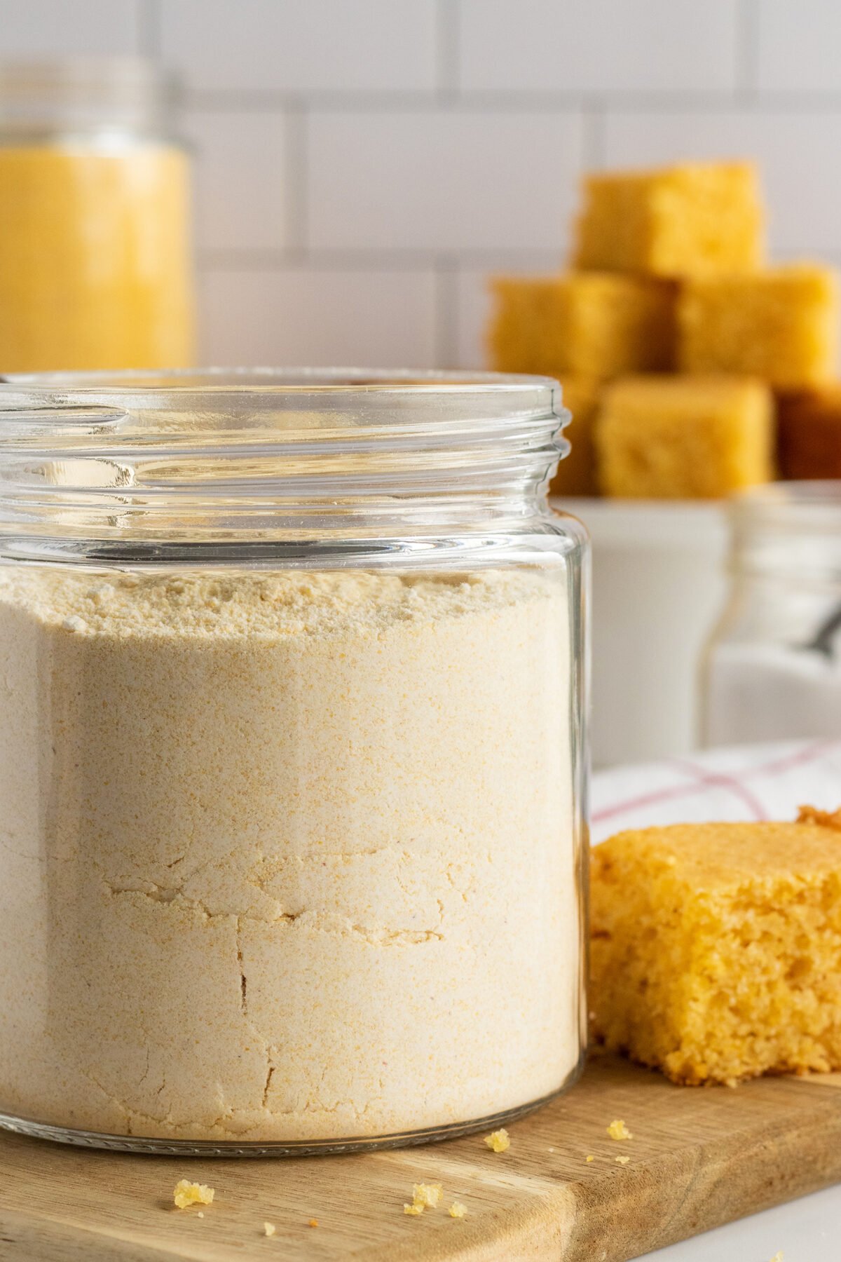 A glass jar filled with cornbread mix sits on a wooden board, with a few pieces of baked cornbread nearby and more cornbread squares stacked in the background. White tiled wall in the background.