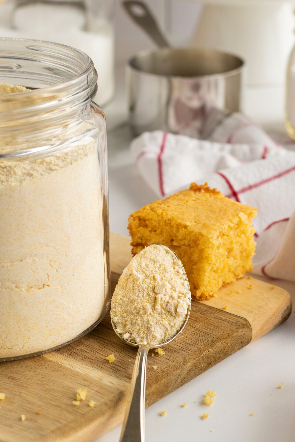 A spoonful of cornmeal rests in front of a glass jar filled with cornmeal, beside a slice of cornbread on a wooden cutting board. A kitchen towel and cooking utensils are visible in the background.