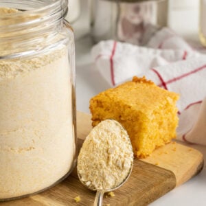 A spoonful of cornmeal rests in front of a glass jar filled with cornmeal, beside a slice of cornbread on a wooden cutting board. A kitchen towel and cooking utensils are visible in the background.