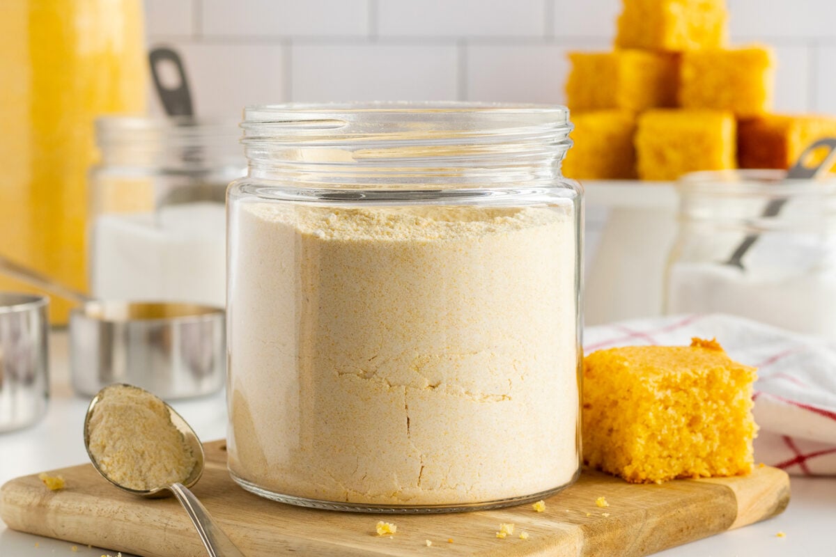 A glass jar filled with cornmeal sits on a wooden board, surrounded by measuring cups, sugar jars, and square pieces of cornbread. A spoon with cornmeal rests in the foreground, and cornbread squares are stacked in the background.