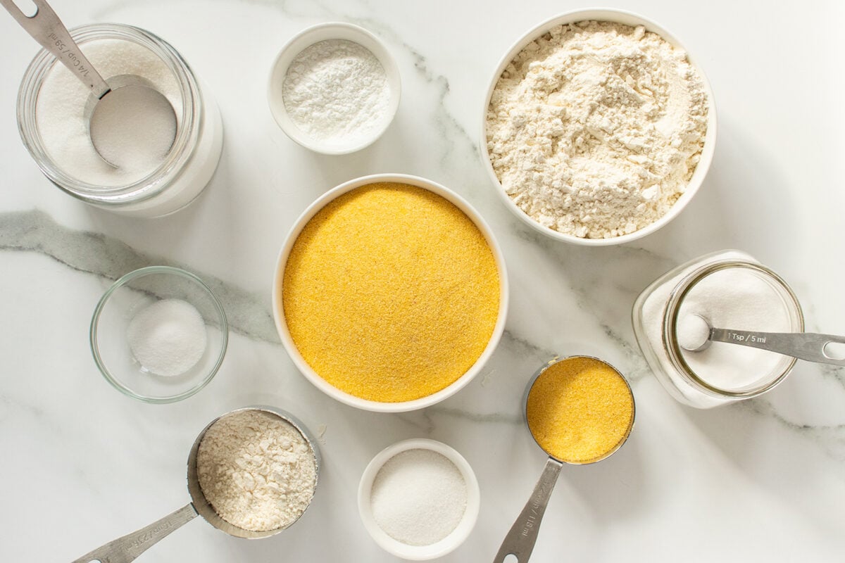 A top-down view of bowls and measuring cups containing cornmeal, flour, sugar, baking powder, and salt, arranged on a white marble surface.