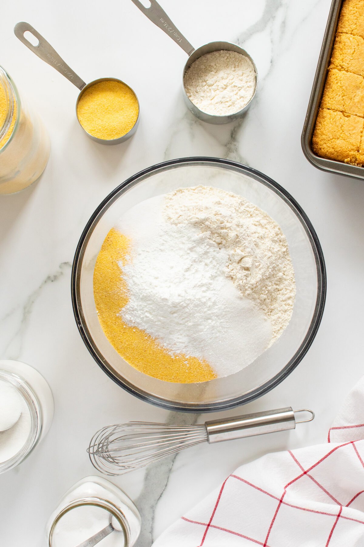 A glass bowl filled with cornmeal, flour, sugar, and baking powder sits on a marble countertop, surrounded by measuring cups, a whisk, a jar of milk, and a baking tray of cornbread.