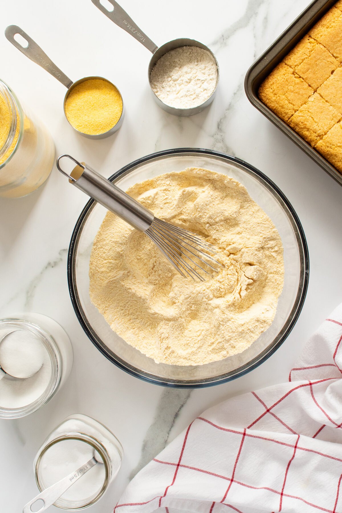 A glass bowl of cornmeal mixture with a whisk, surrounded by measuring cups of flour and cornmeal, a jar of sugar, a baking dish of cornbread, and a white towel with red stripes on a marble countertop.
