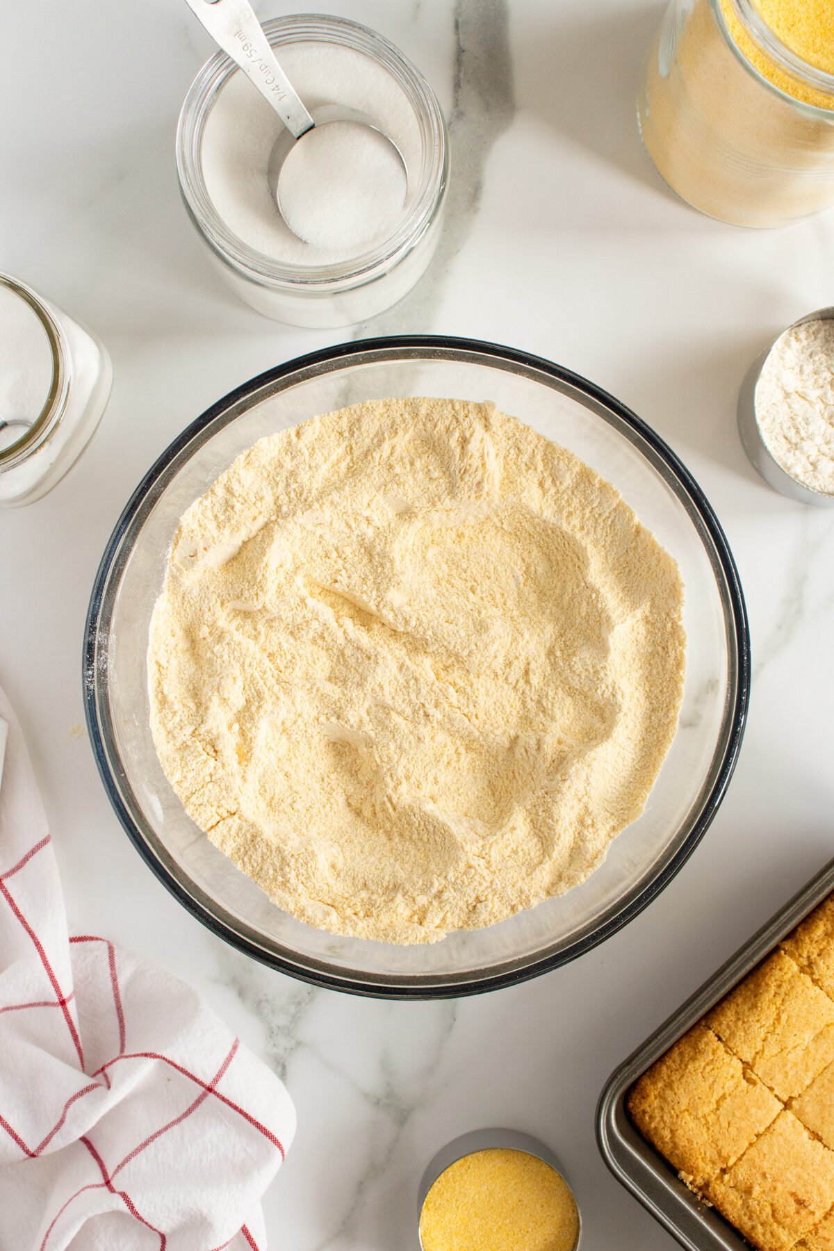 A glass bowl filled with cornmeal sits on a white marble surface, surrounded by baking ingredients like sugar, flour, and salt, with a baked cornbread in a pan nearby and a red-striped towel to the side.