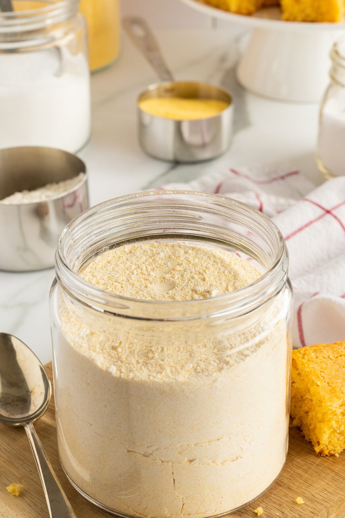 A glass jar filled with cornmeal sits on a wooden surface next to a spoon, with cornbread, measuring cups, and a jar of flour in the background.
