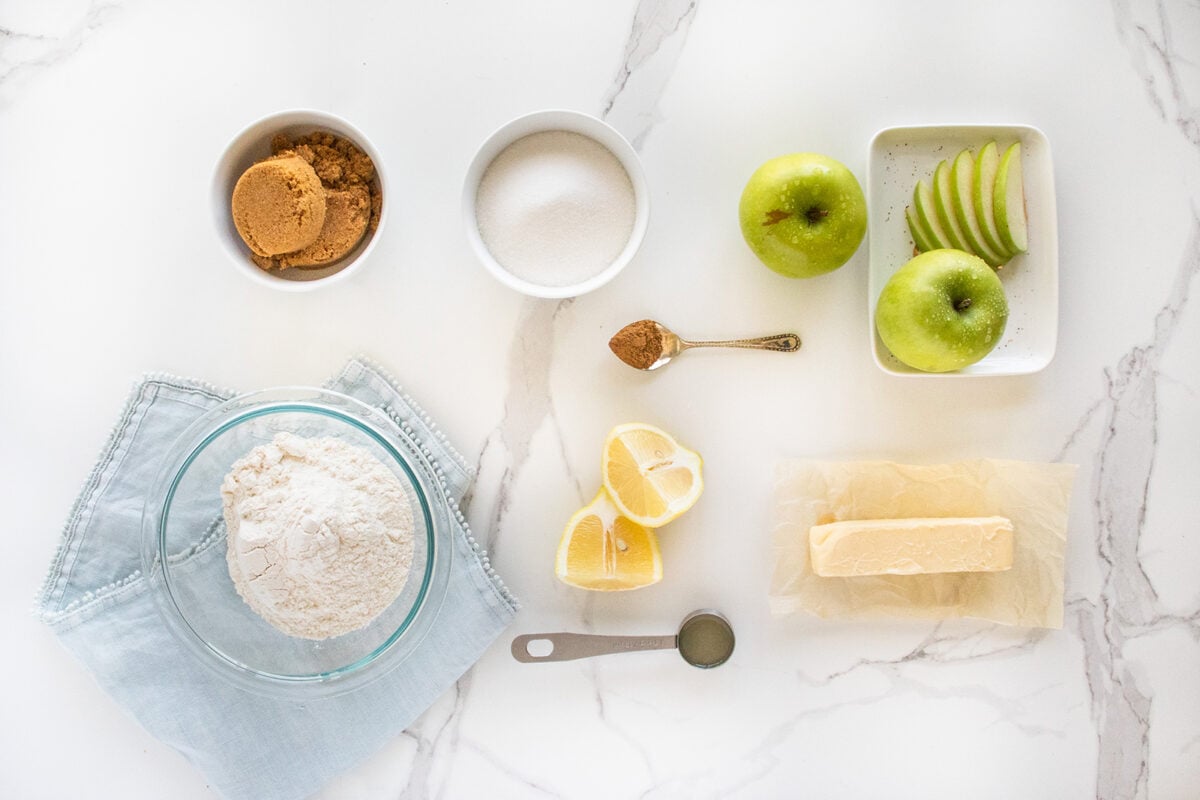 Ingredients for baking: a bowl of flour, a bowl of sugar, brown sugar, two green apples (one sliced), a lemon cut in wedges, a stick of butter, a spoon of cinnamon, and a spoon of vanilla on a white surface.