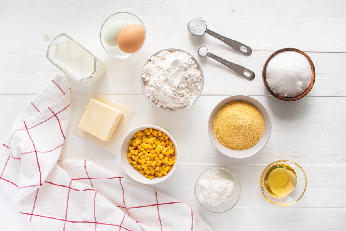 A flat lay of cornbread ingredients on a white surface: flour, cornmeal, sugar, sour cream, milk, butter, oil, two eggs, corn kernels, measuring spoons, and a white towel with red stripes.