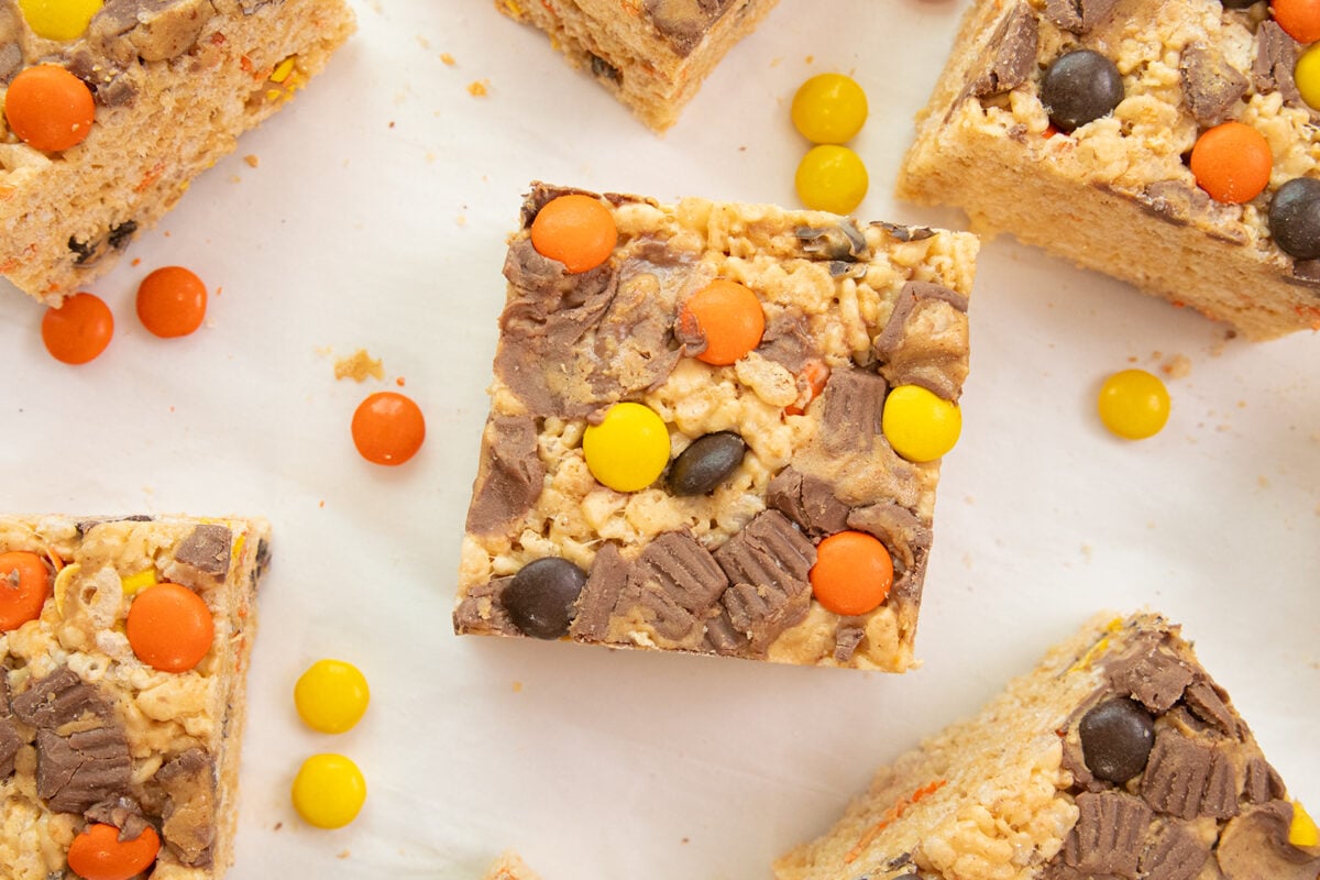 A close-up of rice crispy treat squares topped with chocolate and colorful candy-coated chocolates, arranged on a white surface with a few candies scattered around.