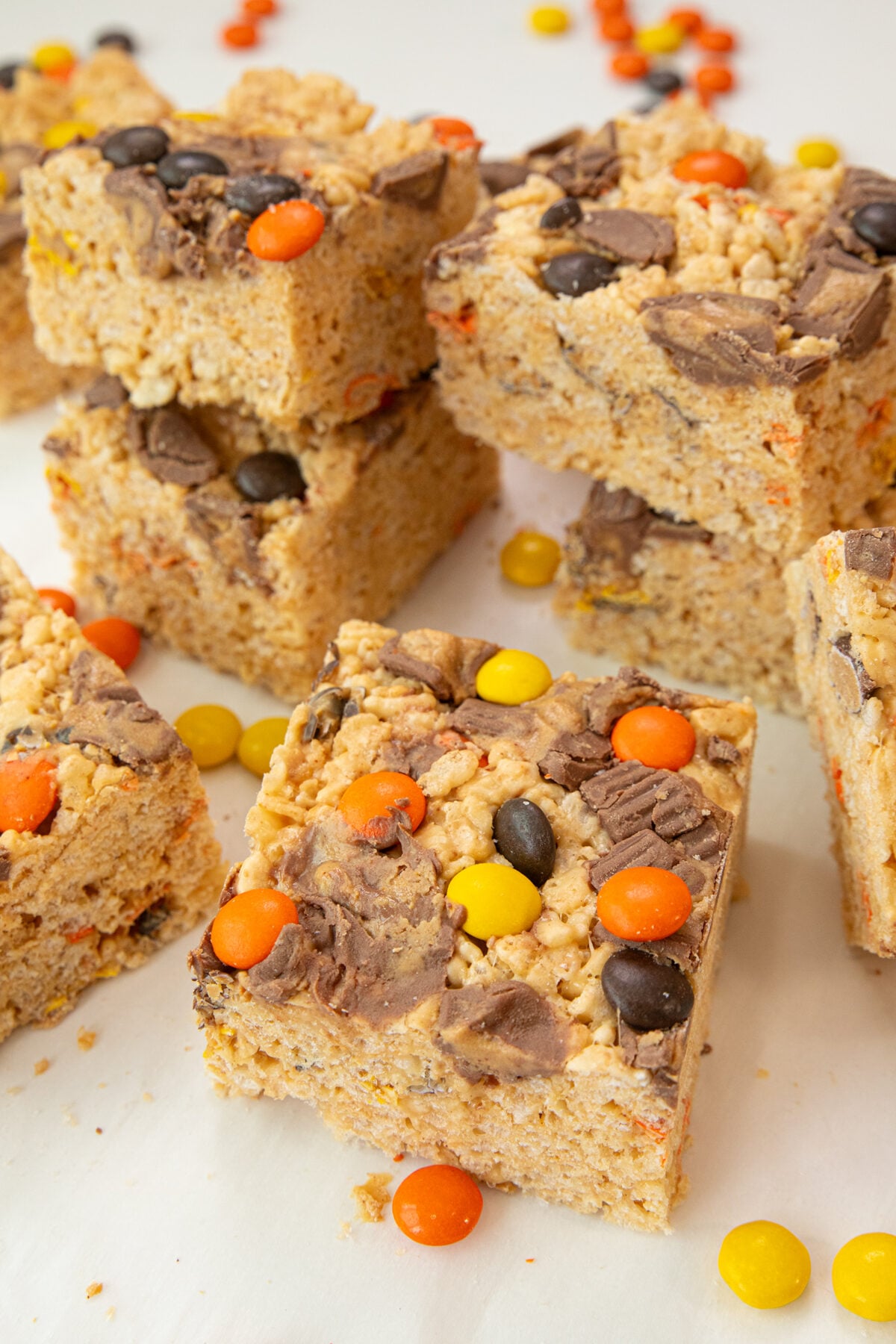 Rice Krispies treats cut into squares, topped with chopped chocolate and orange, yellow, and brown candy-coated pieces, are stacked on a white surface. Some loose candies are scattered around.