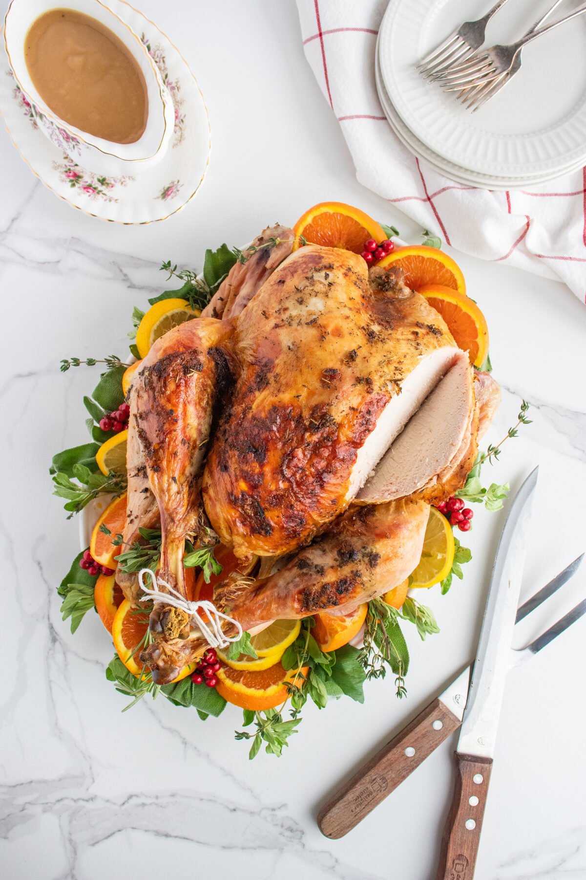 A roasted turkey garnished with orange slices, herbs, and cranberries sits on a platter. A carving set, gravy boat, and stacked white plates with forks are nearby on a white marble surface.