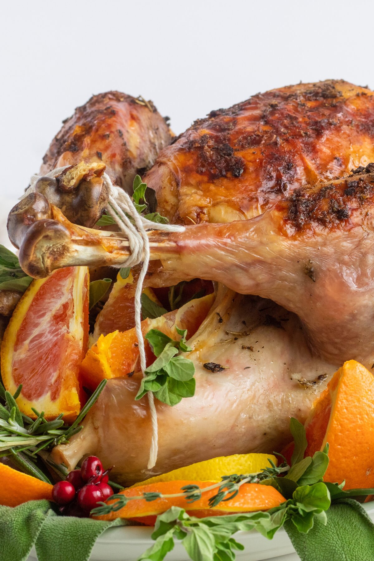 A roasted turkey garnished with fresh herbs, orange slices, and cranberries is displayed on a platter against a white background. The turkey has crispy, browned skin and is surrounded by vibrant, fresh toppings.