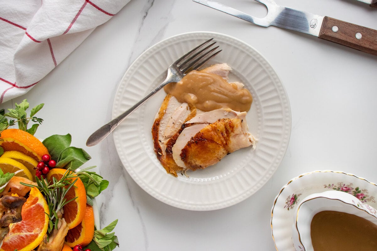 A white plate with sliced roasted turkey and brown gravy, topped with a fork. Next to it is a platter of herbs, oranges, and cranberries, and a carving fork on a white marble surface.