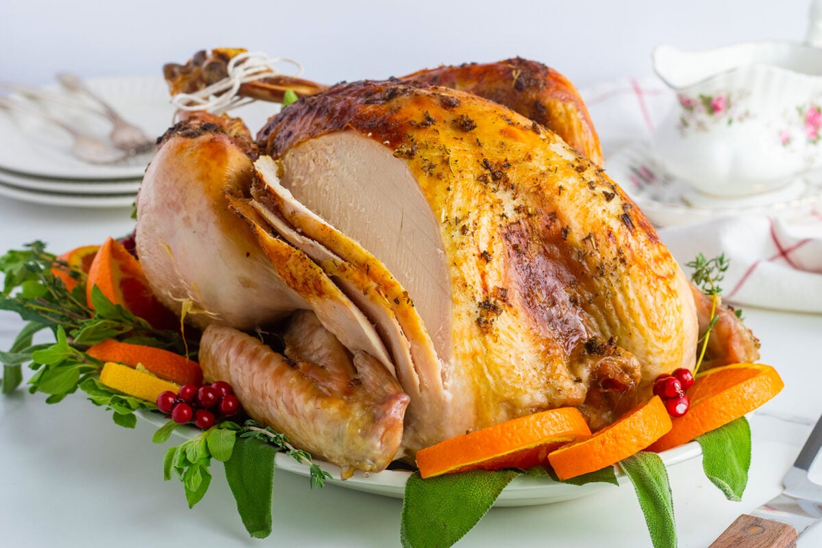A golden-brown roasted turkey sits on a platter, garnished with orange slices, cranberries, herbs, and greens. Plates, utensils, and a floral-patterned gravy boat are in the background on a white table.