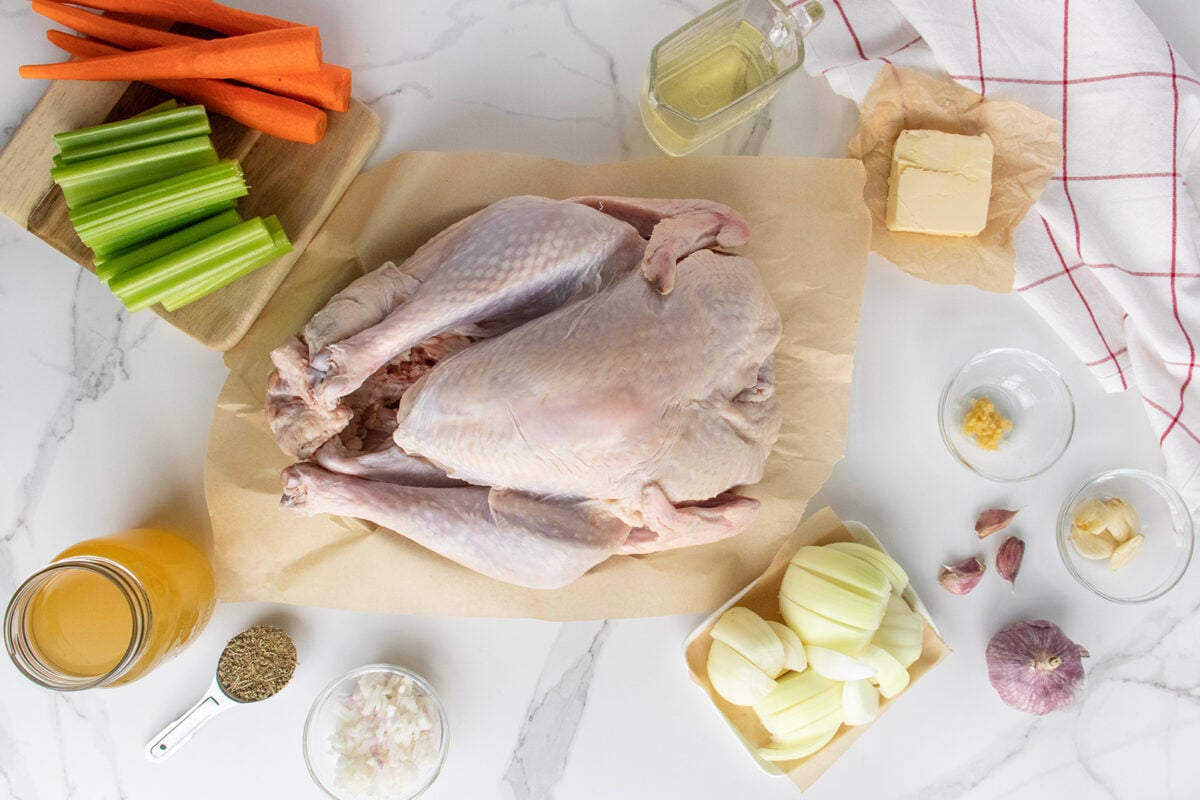 Raw turkey on parchment paper is surrounded by celery, carrots, onion, garlic, butter, oil, broth, shallots, thyme, and a red-checked towel on a white marble countertop, arranged neatly for meal preparation.