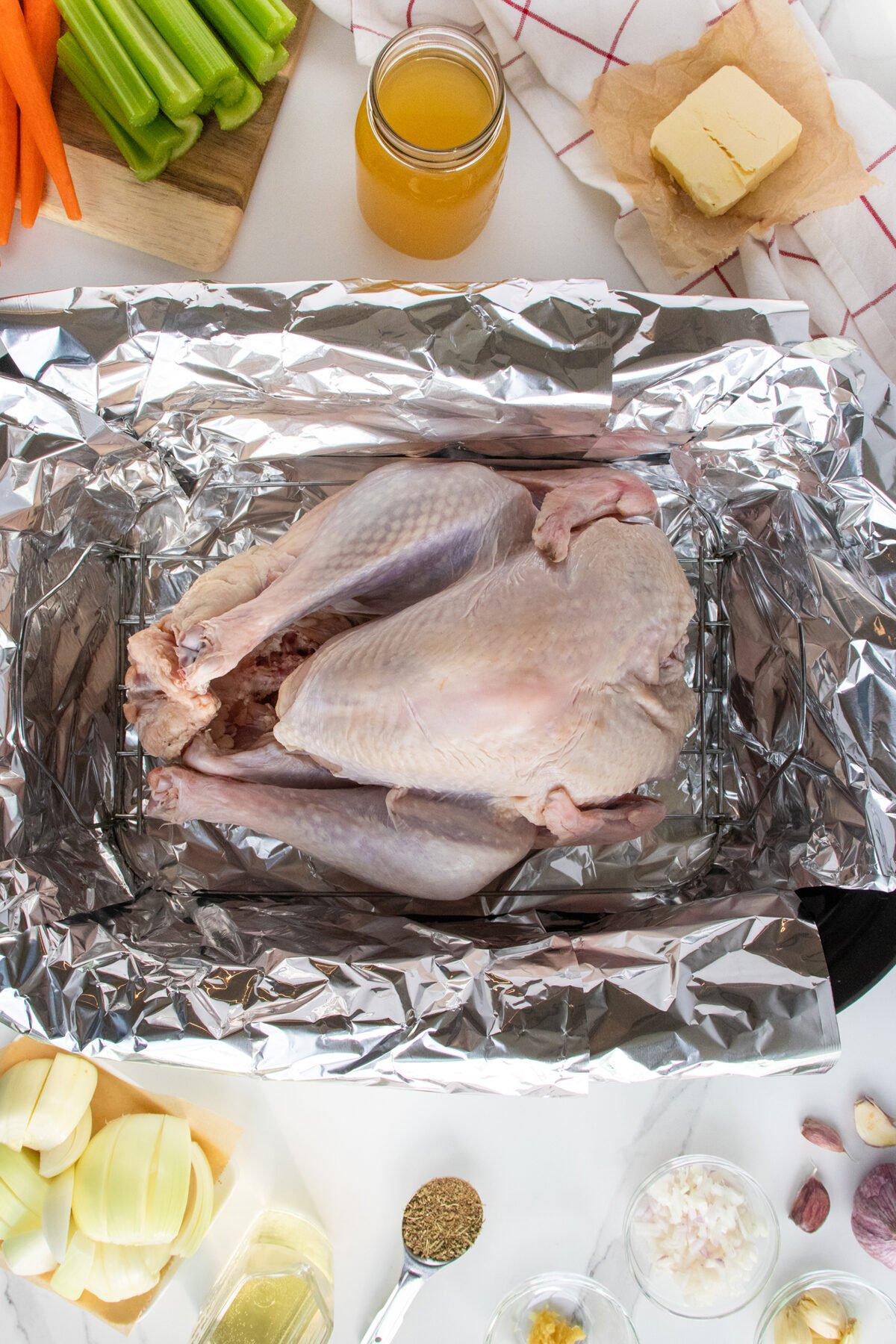 A raw whole turkey sits in a foil-lined roasting pan, surrounded by ingredients including celery, carrots, onion, broth, butter, oil, herbs, and garlic on a white countertop.