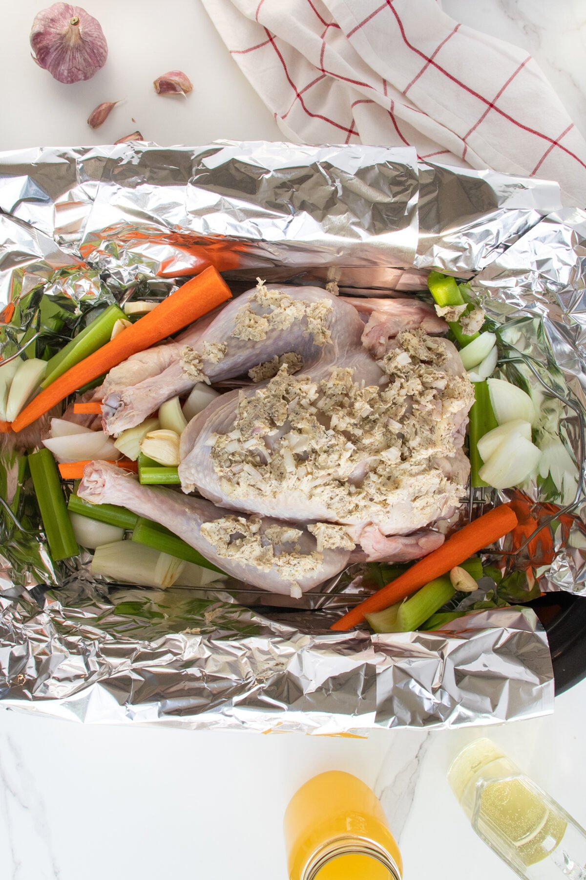 Raw, seasoned chicken coated with herbs rests on chopped carrots, celery, and onions in a foil-lined baking tray, with garlic cloves and a kitchen towel nearby on a marble countertop.