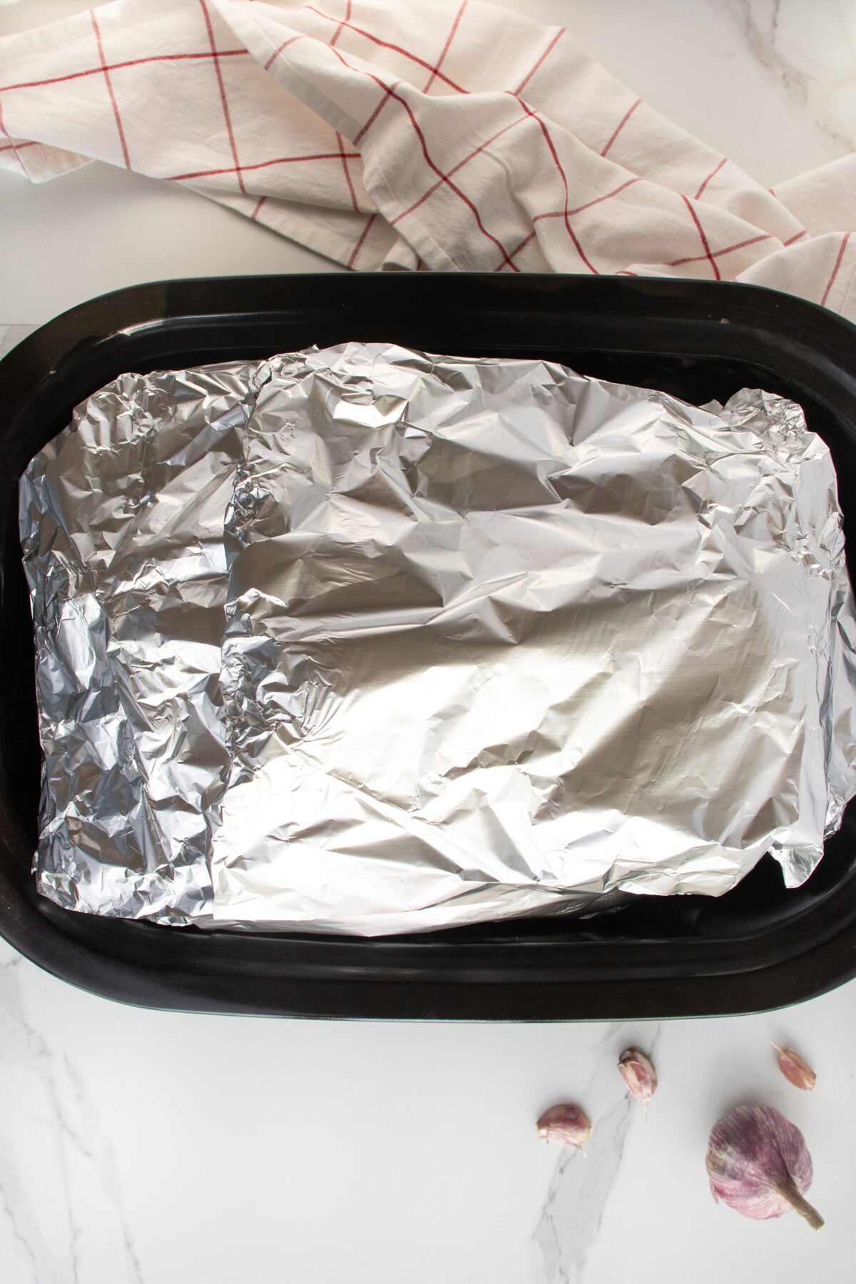 A black roasting pan with food covered in aluminum foil, placed on a white marble surface. A white and red checkered towel is in the background, and garlic cloves are nearby.