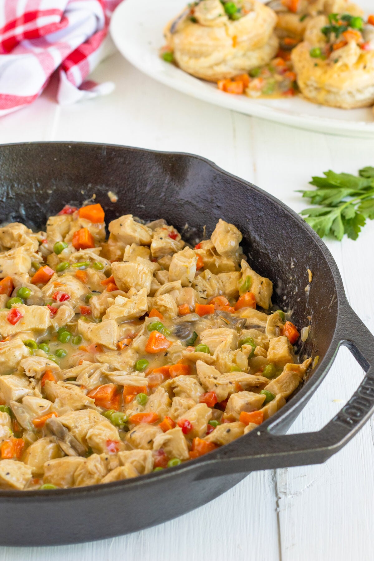 A cast iron skillet filled with creamy chicken pot pie filling, including diced chicken, peas, carrots, and mushrooms. In the background, biscuit-topped pot pies rest on a white plate.
