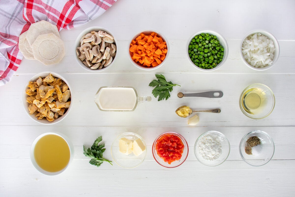 Overhead view of bowls with chopped mushrooms, carrots, peas, onion, cooked chicken, broth, cream, butter, flour, diced tomatoes, oil, garlic, parsley, and seasonings on a white table with a red checkered cloth.