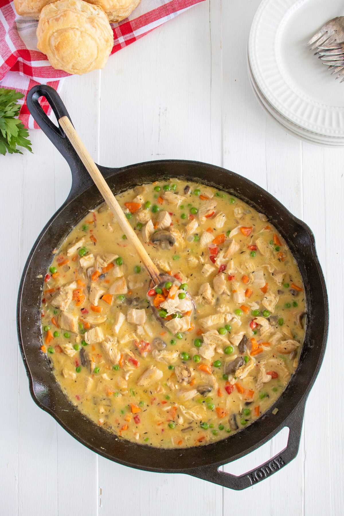 A cast iron skillet filled with creamy chicken and vegetable stew, including peas, carrots, and mushrooms, sits on a white wooden table. A wooden spoon rests inside the skillet. Biscuits and plates are nearby.