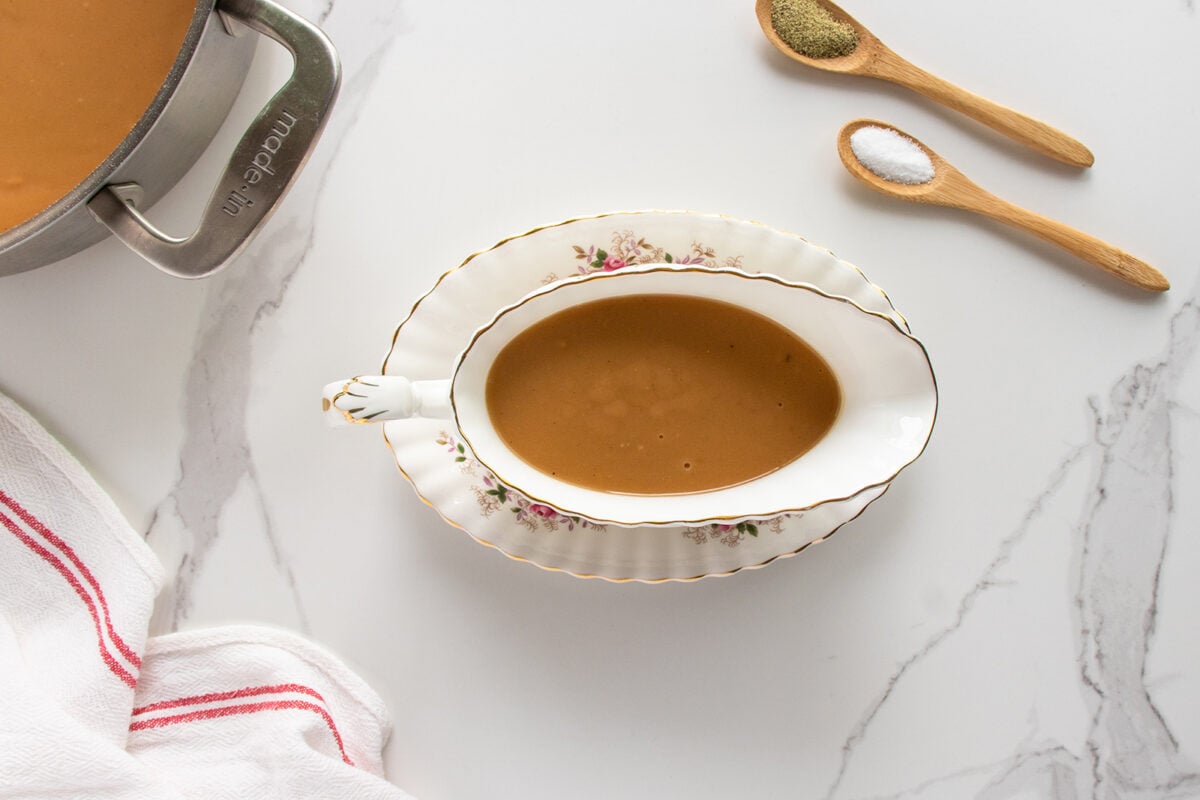 A white gravy boat filled with brown gravy sits on a marble surface beside a pot, a towel, and two wooden spoons holding salt and pepper.