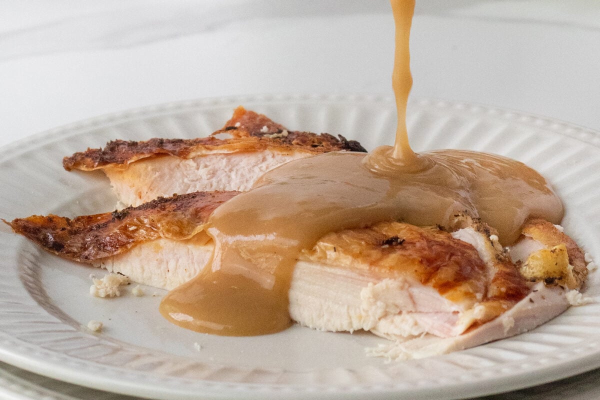 Slices of roasted turkey breast with crispy skin on a white plate, topped with brown gravy being poured over the meat.