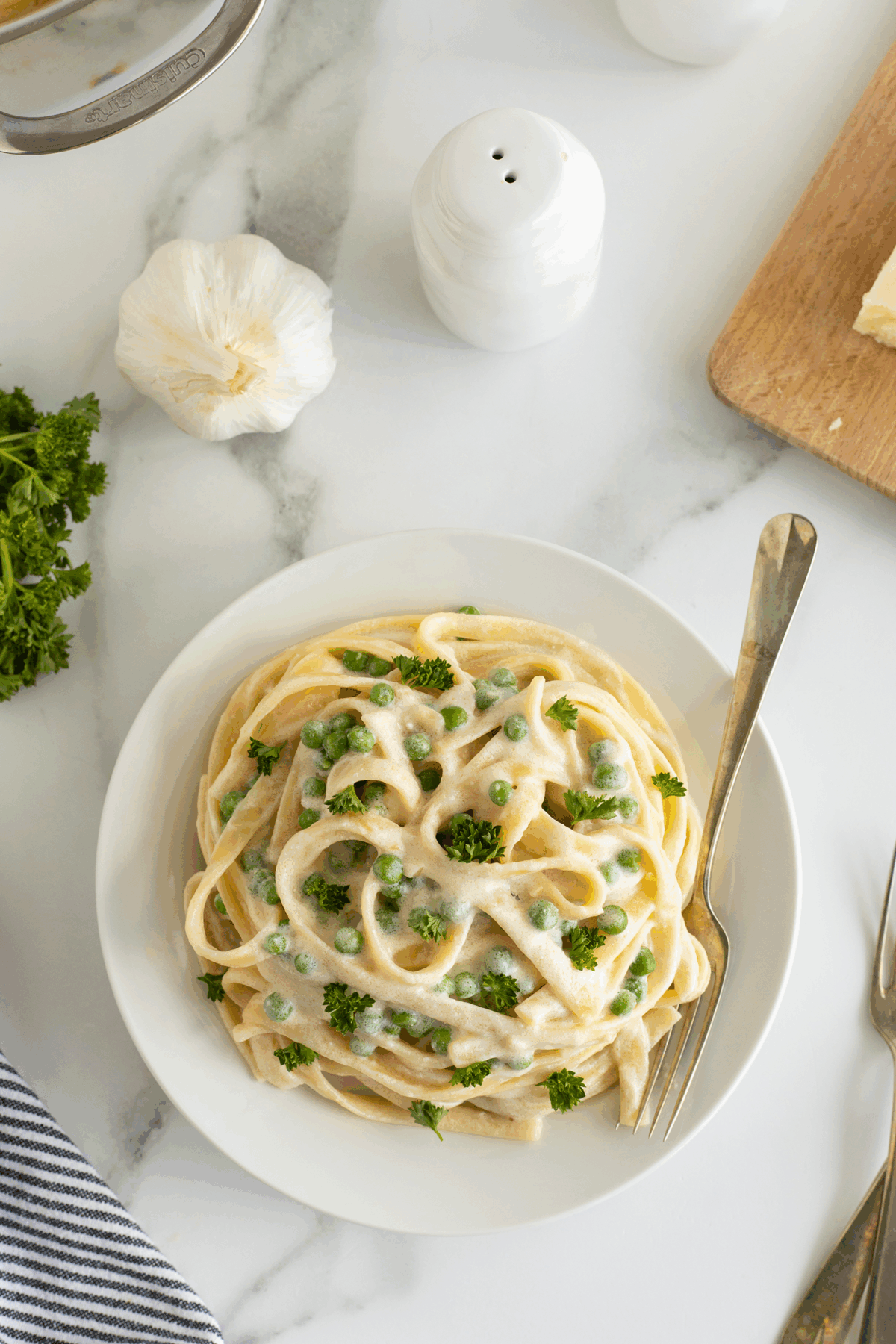 A bowl of fettuccine pasta with creamy white sauce, green peas, and parsley garnish sits on a white marble surface, with garlic, parsley, a pepper shaker, and a fork nearby.