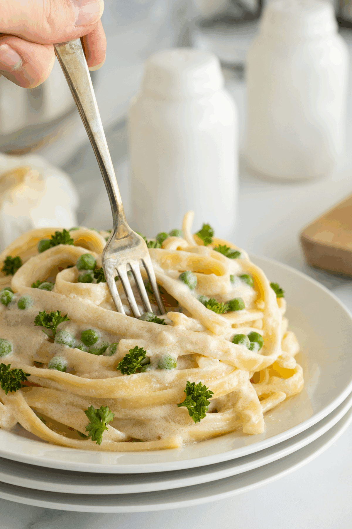 A hand twirls a fork in creamy pasta topped with green peas and parsley, served on a white plate stacked on another plate. Salt and pepper shakers are blurred in the background.