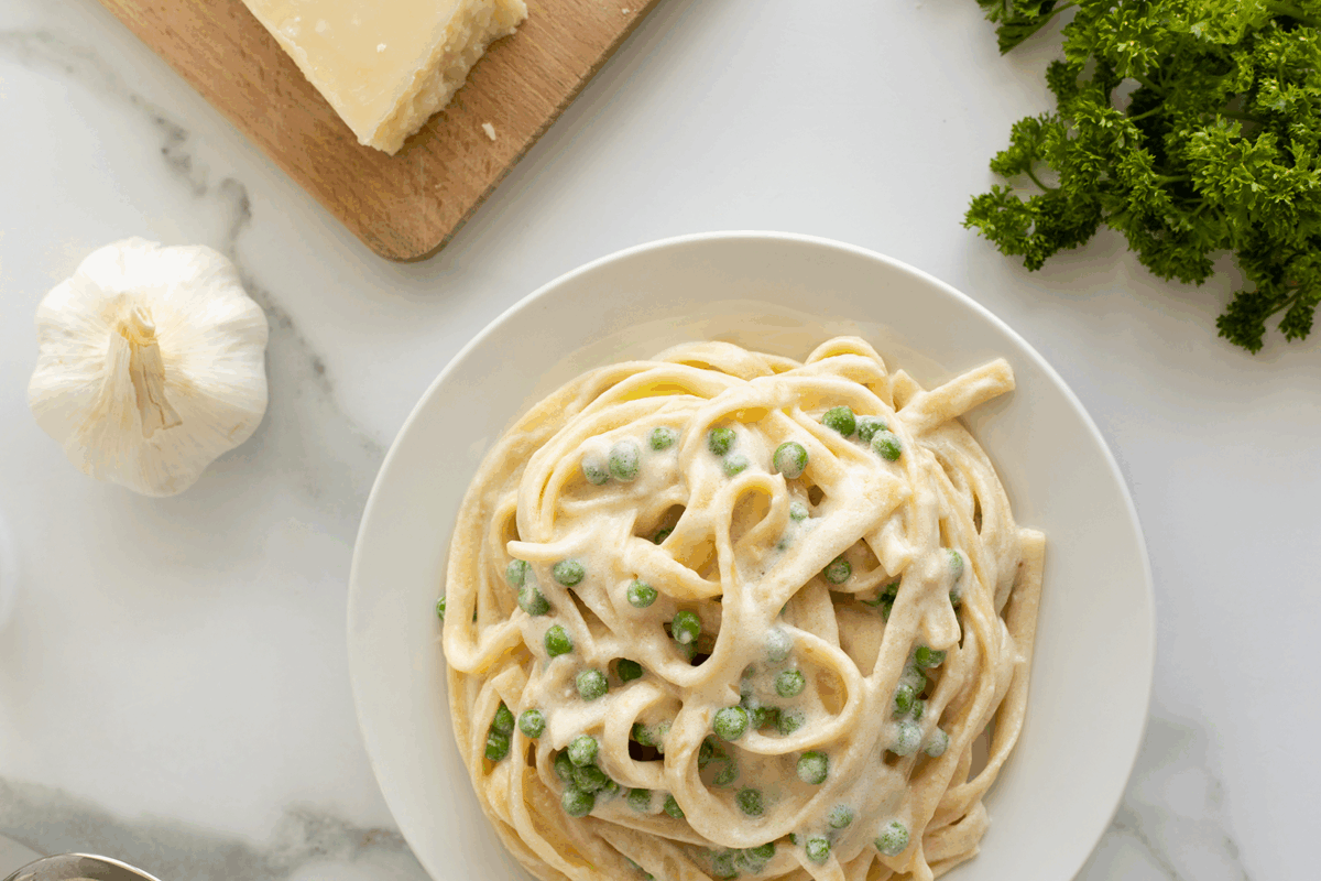 A white plate of fettuccine pasta with creamy sauce and green peas sits on a marble surface, surrounded by a garlic bulb, a block of cheese on a wooden board, and fresh parsley.