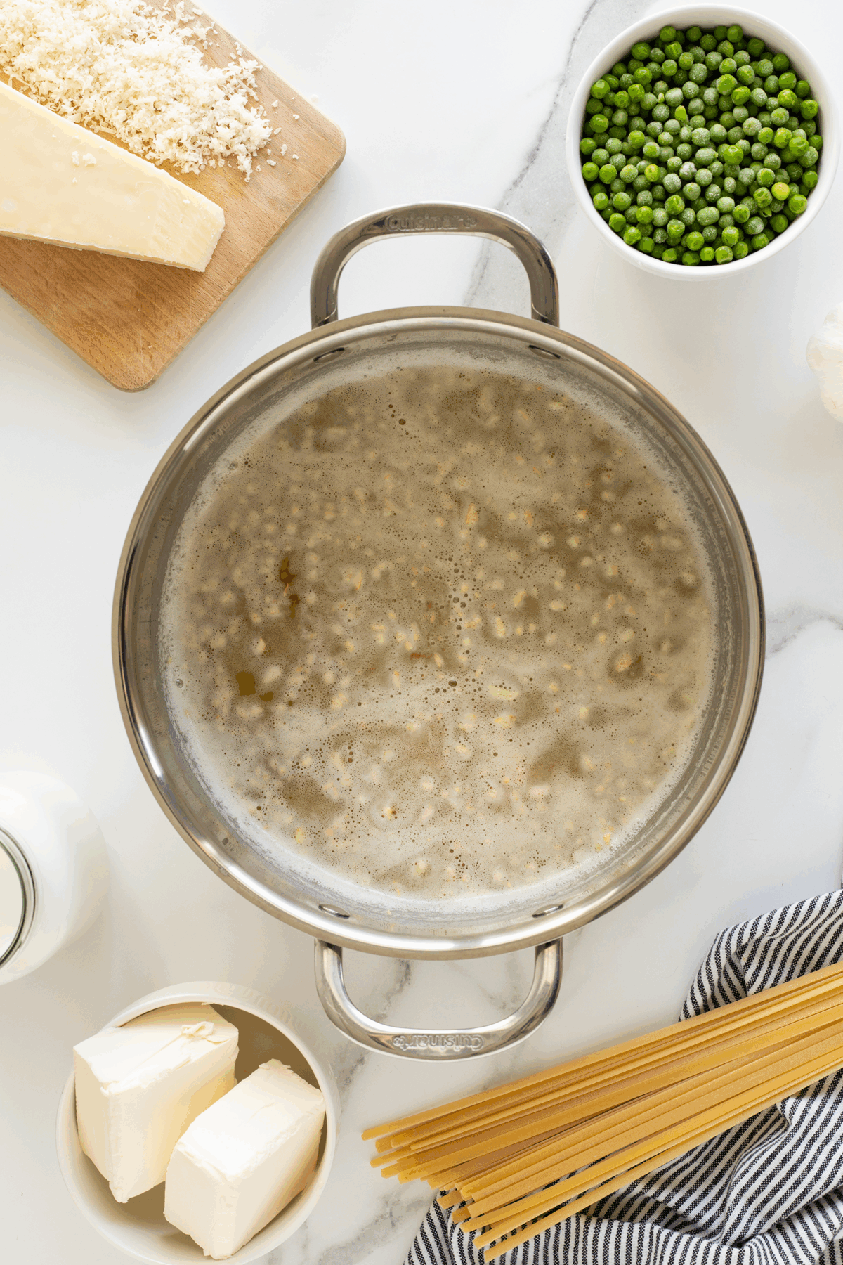 A pot of boiling water with pasta cooking inside, surrounded by parmesan cheese, frozen peas, butter, uncooked spaghetti, and a bottle of milk on a white countertop.