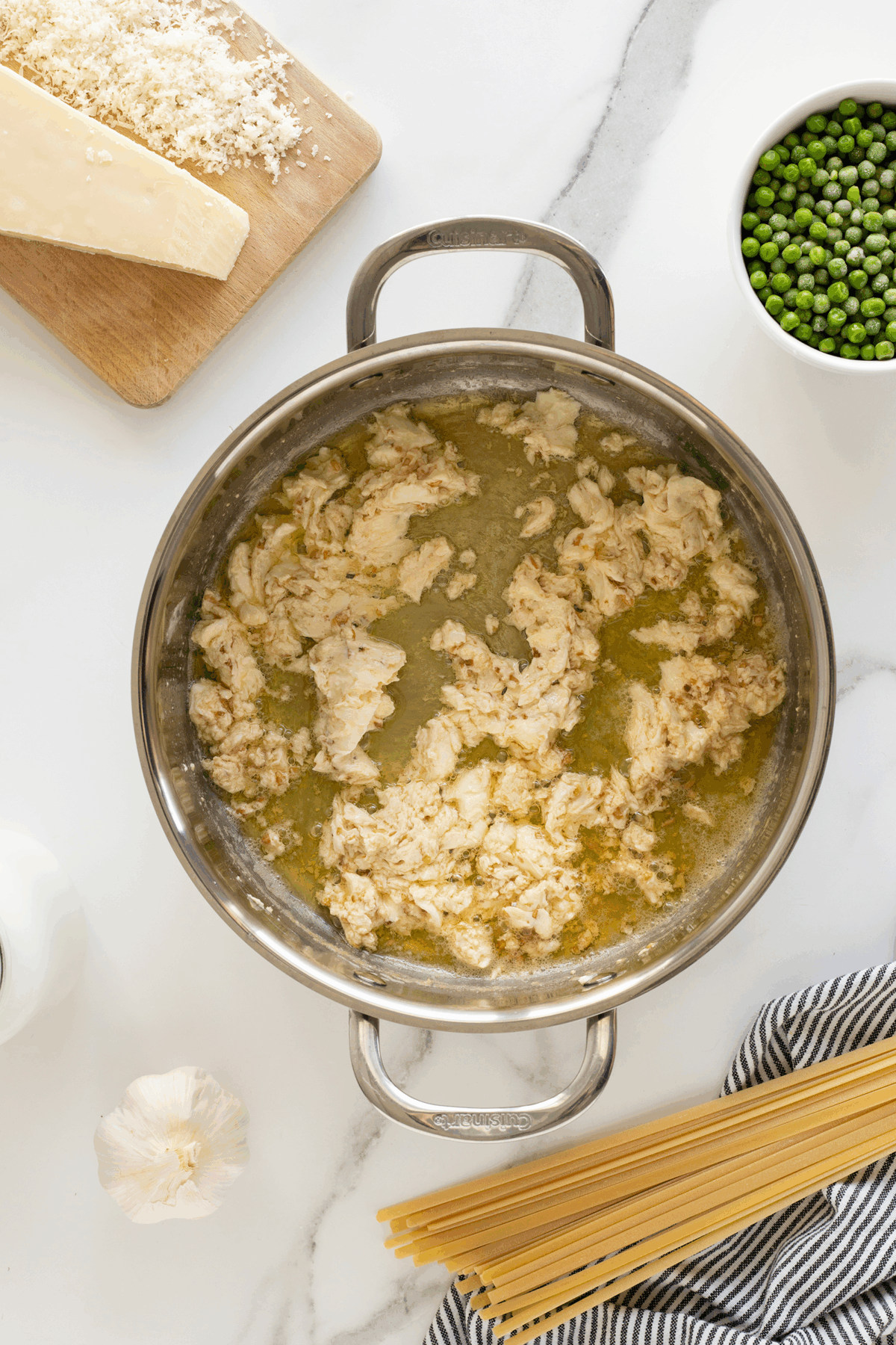 A pot of cooked ground chicken in broth sits on a marble countertop, surrounded by a block of cheese, shredded cheese, a bowl of green peas, uncooked spaghetti, a garlic bulb, and a bottle of milk.