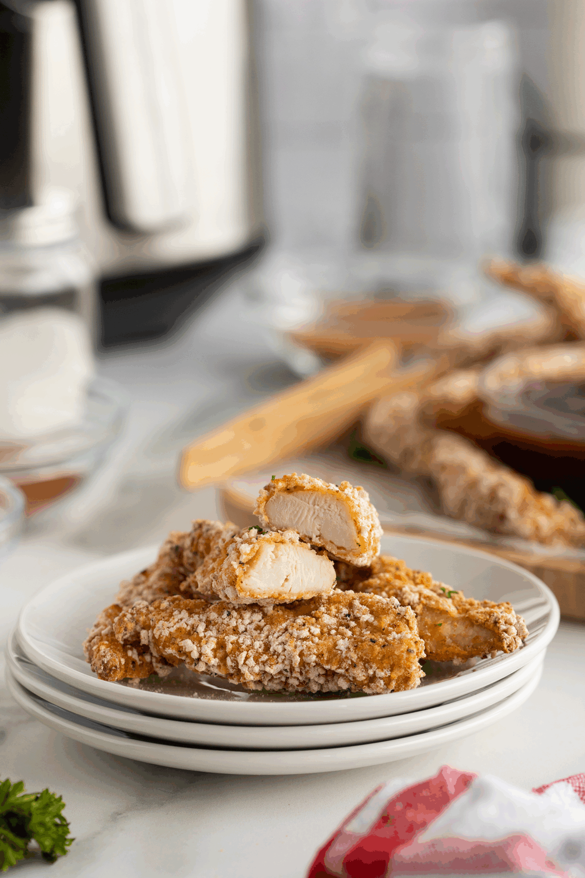 A stack of breaded chicken tenders sits on a white plate, with one piece cut open to show the juicy interior. The background features blurred kitchen items and more chicken tenders on a wooden tray.