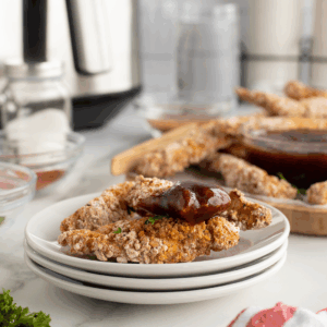 Three breaded chicken tenders on a white plate, topped with barbecue sauce, with more tenders and a bowl of sauce in the background. Parsley and seasonings are on the countertop nearby.