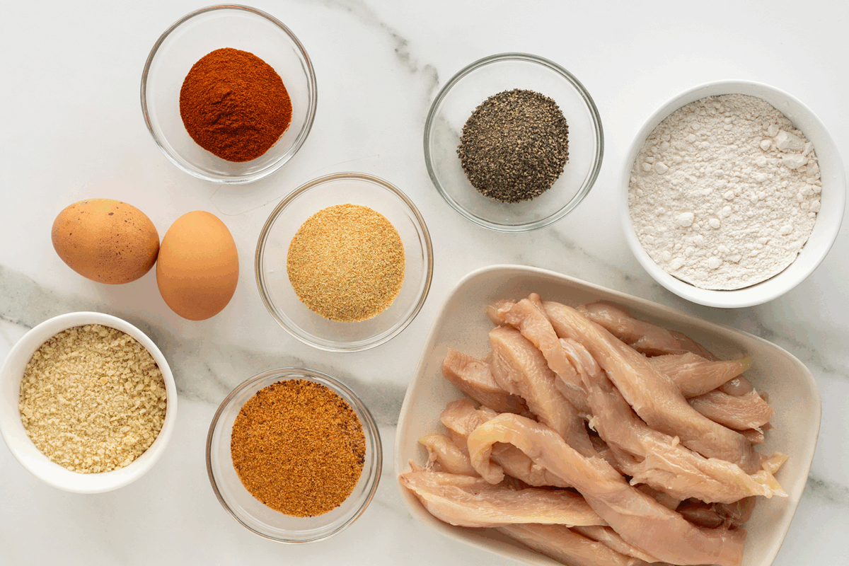 Raw chicken strips, two eggs, flour, black pepper, paprika, bread crumbs, seasoned breading, and garlic powder are arranged in small bowls on a white marble surface.