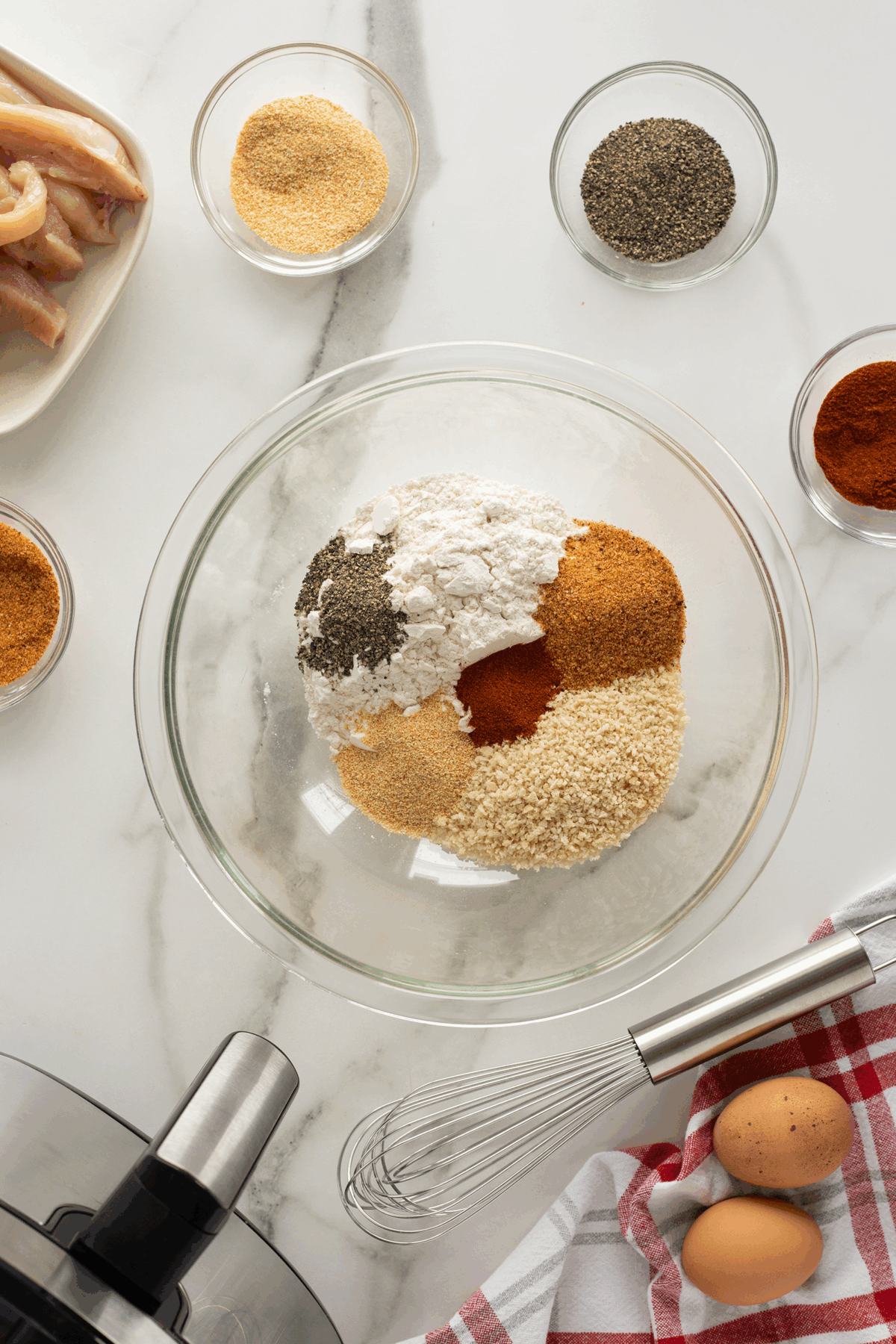 A glass bowl with flour, breadcrumbs, and assorted spices sits on a marble countertop, surrounded by a whisk, two eggs, a red-checked towel, and small bowls of seasonings. Sliced raw chicken is nearby.