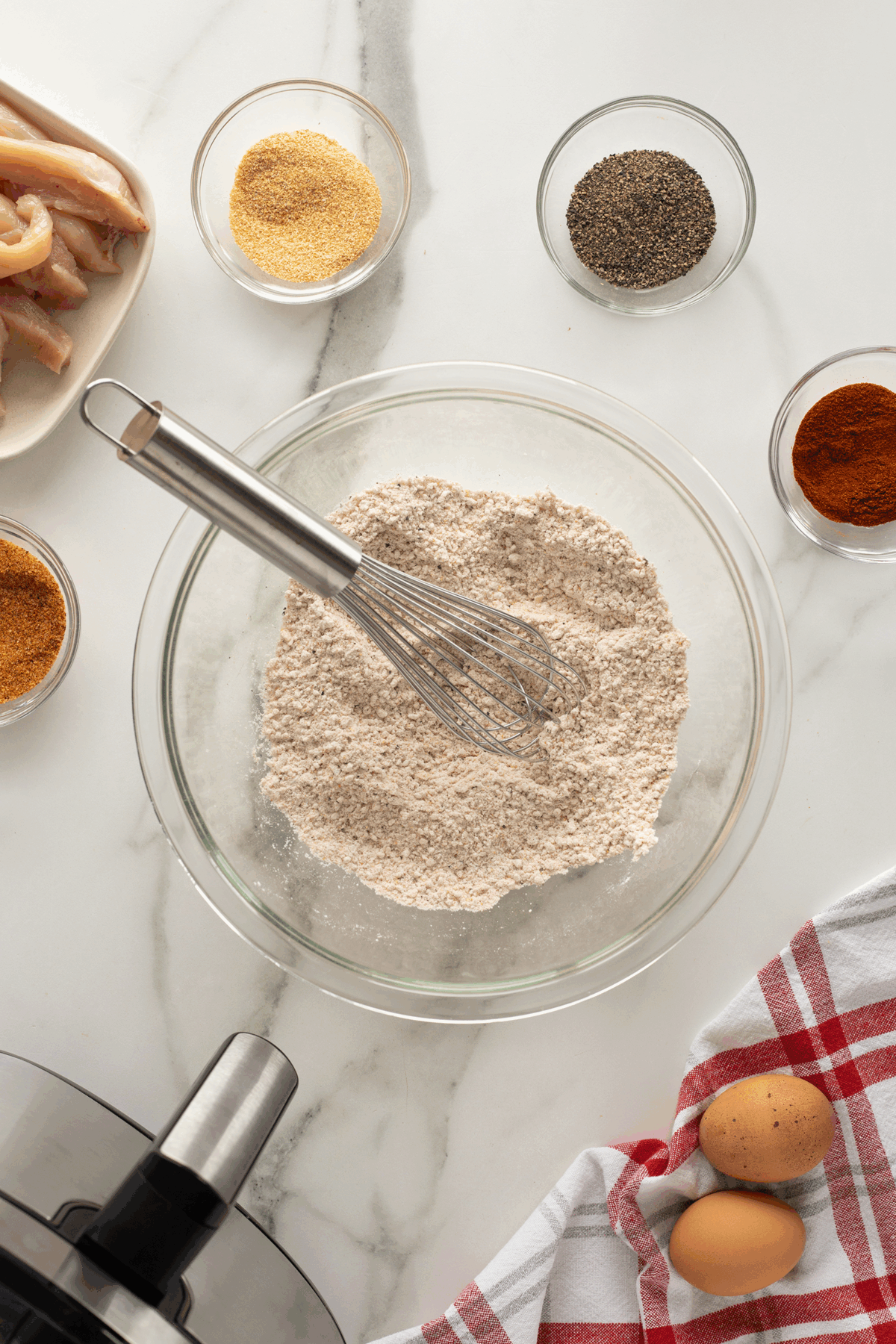 A bowl of flour with a whisk sits on a marble counter surrounded by bowls of spices, raw chicken strips, two eggs on a red-and-white towel, and part of an air fryer in the corner.
