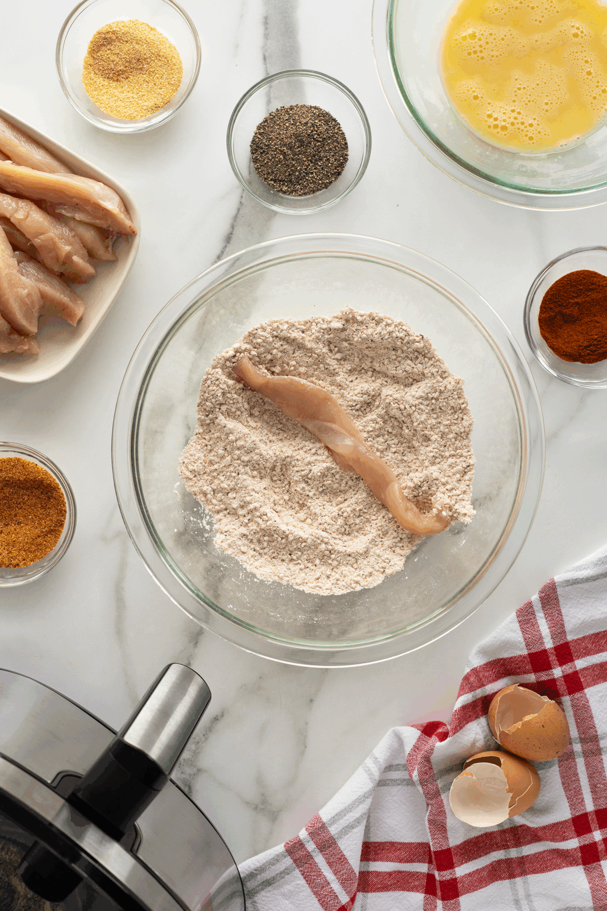 A raw chicken strip being coated in flour mixture in a glass bowl, surrounded by bowls of spices, egg wash, breadcrumbs, a plate of chicken strips, and broken eggshells on a red and white towel on a marble countertop.