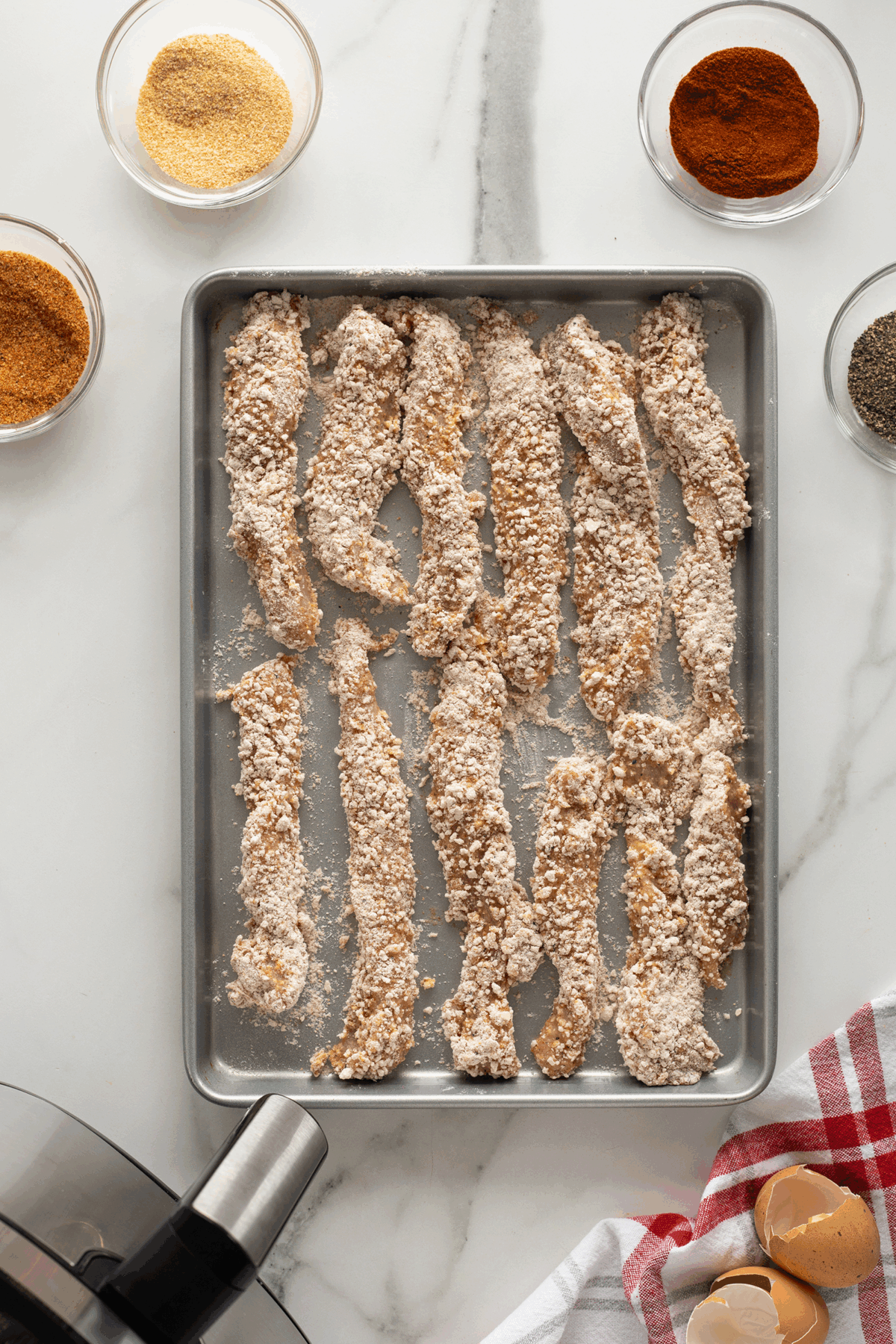 A baking tray with uncooked, breaded chicken strips sits on a marble surface, surrounded by bowls of spices and breadcrumbs, cracked eggs, a red-checkered towel, and a food processor base.