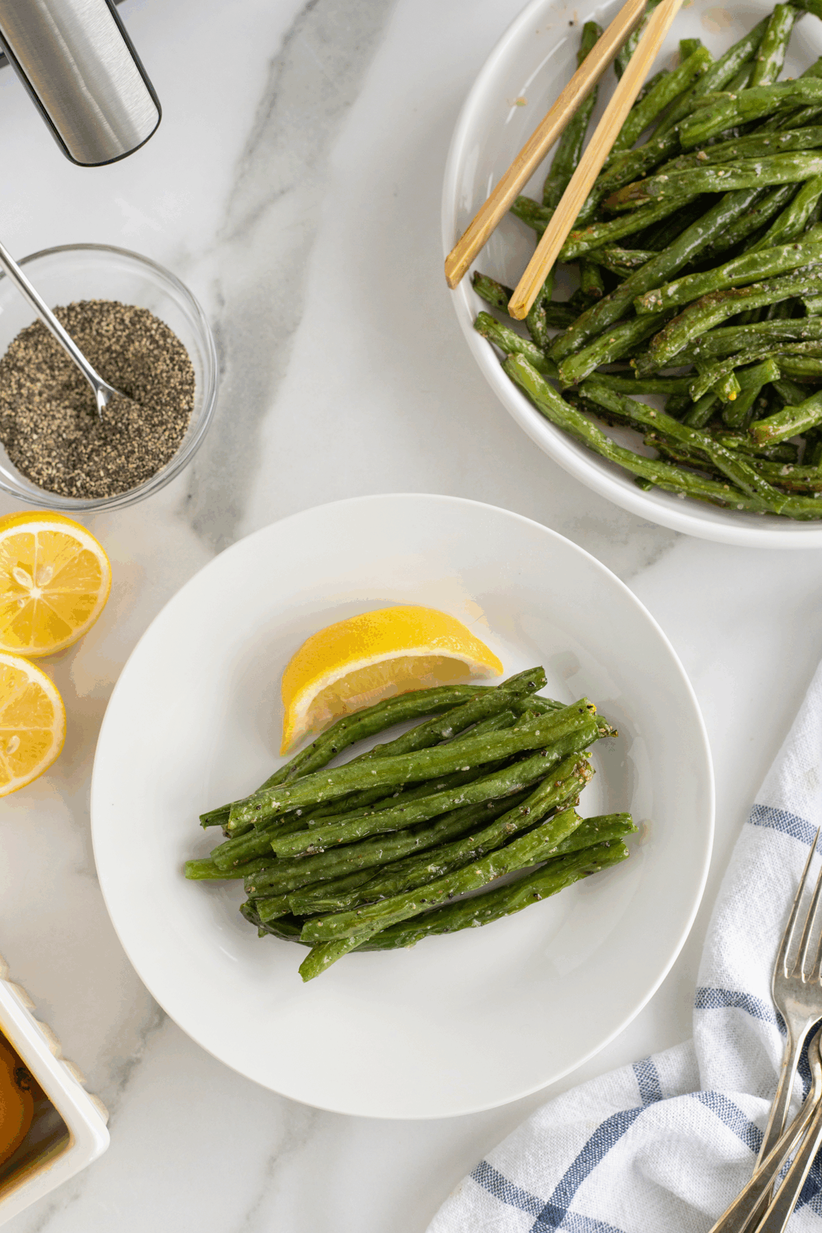 A white plate with roasted green beans and a lemon wedge, next to a bowl of more green beans with chopsticks, a halved lemon, pepper, and a blue-striped napkin on a marble surface.